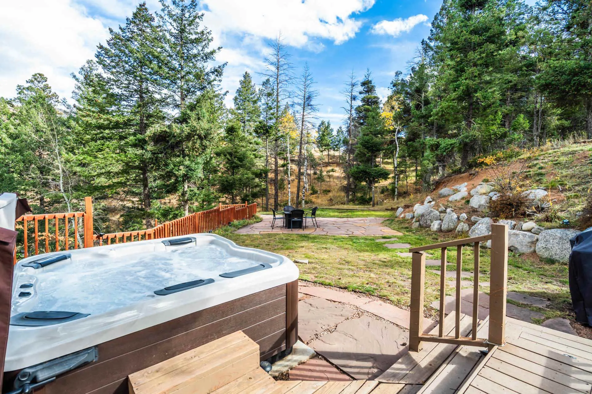 A hot tub sits on a deck next to a wooded hillside, with a patio and table in the yard.