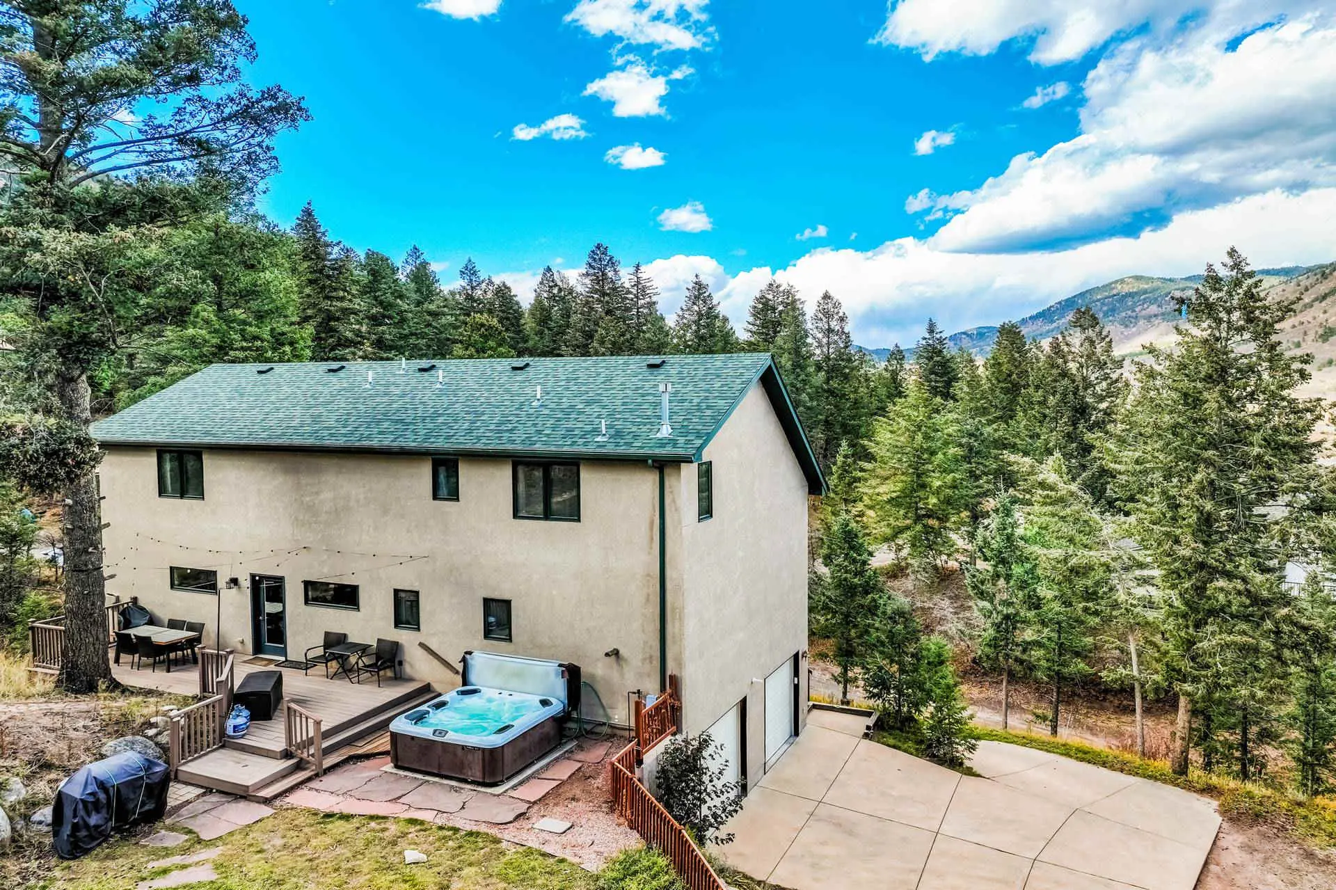 A stucco house with a green roof sits in a wooded area, featuring a hot tub and a deck.