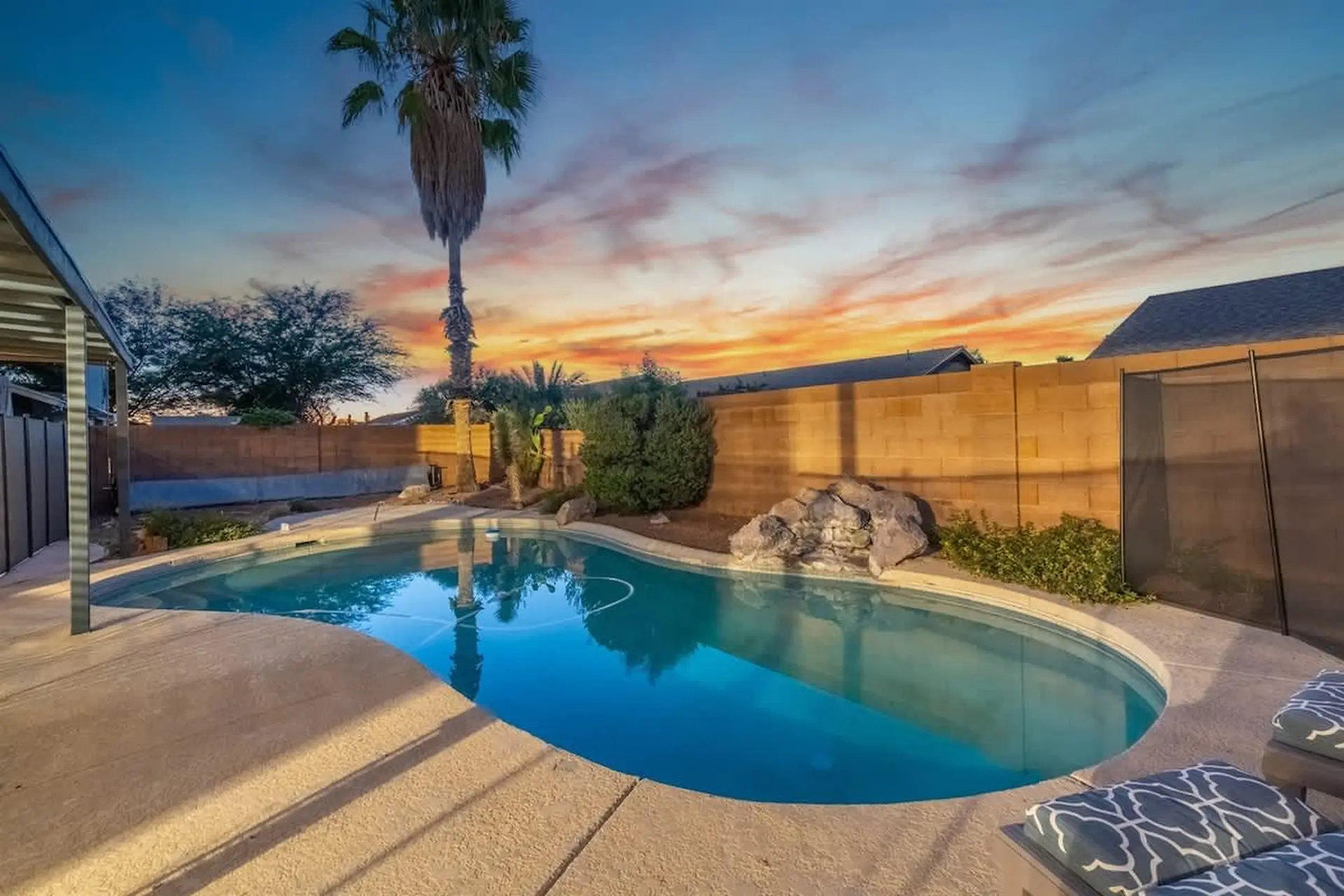 A backyard pool at sunset with a palm tree, rock waterfall, and lounge chairs.