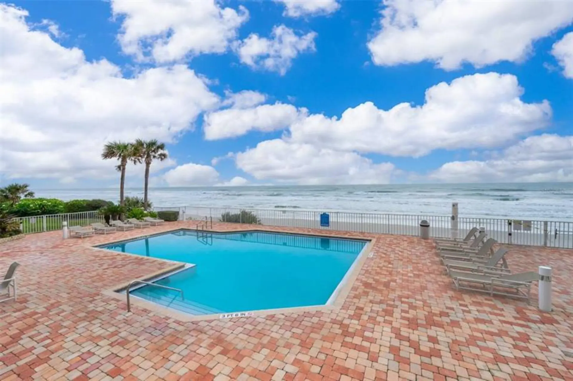 A bright blue swimming pool is surrounded by red brick pavement and overlooks the ocean and palm tre
