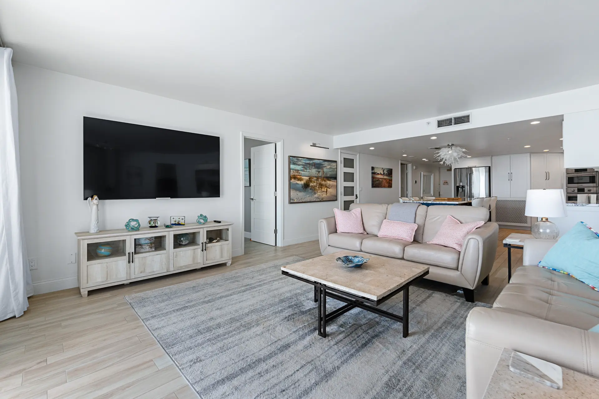 A bright and spacious living room features a tan sofa, cream furniture, and a large TV.