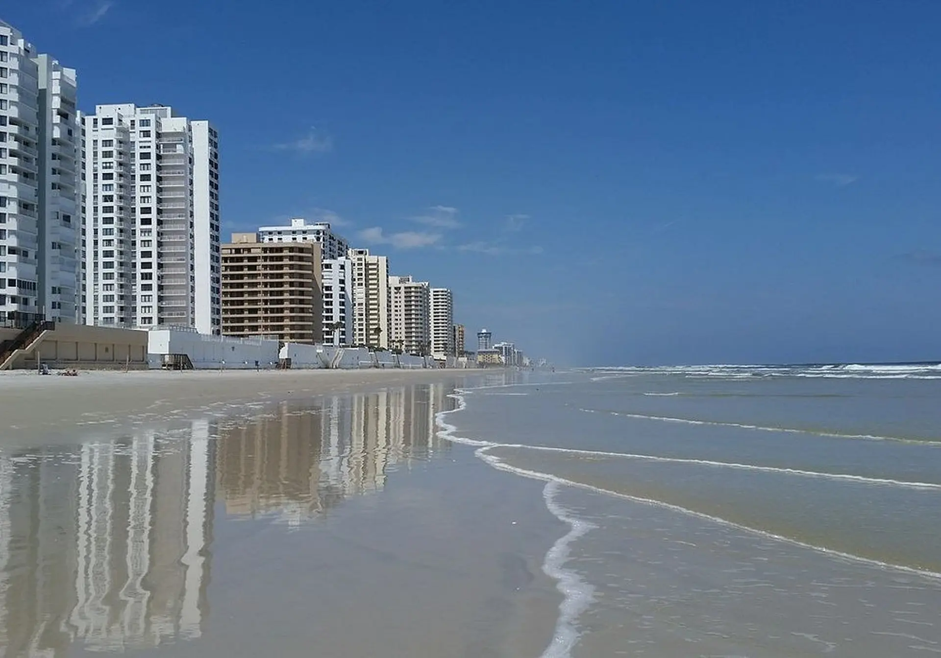 A row of tall white buildings stands on a sandy beach, reflected in the wet sand.