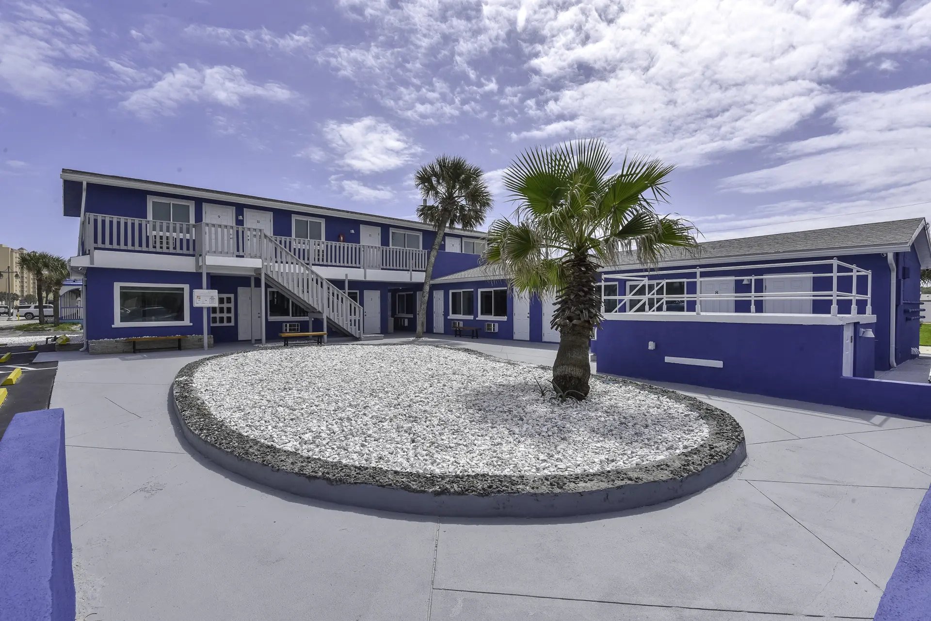 A blue motel with white trim and stairs, a central gravel bed with palm trees, and a cloudy sky.