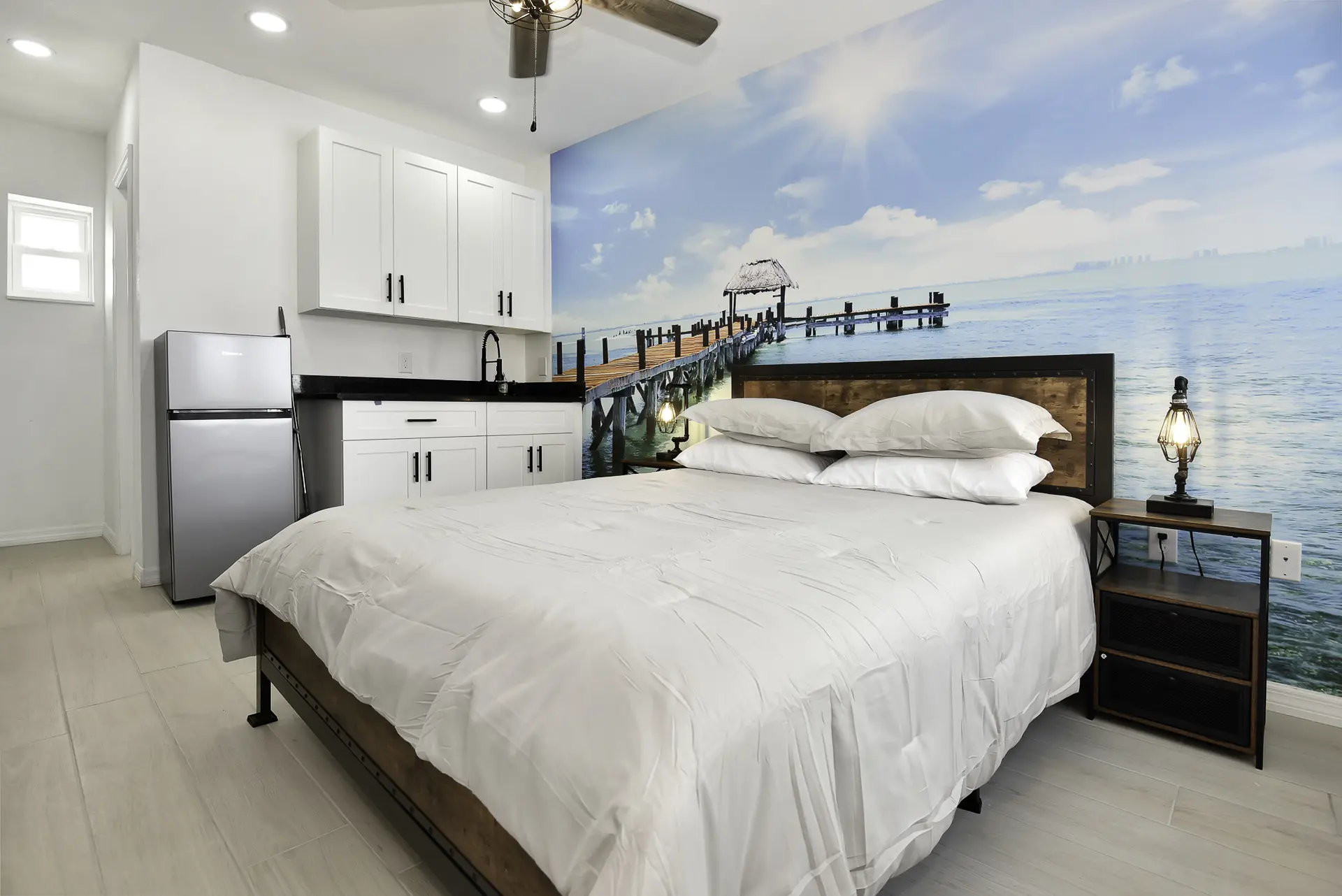 Bedroom with a mural of a pier and ocean, queen bed, kitchen cabinets and refrigerator.