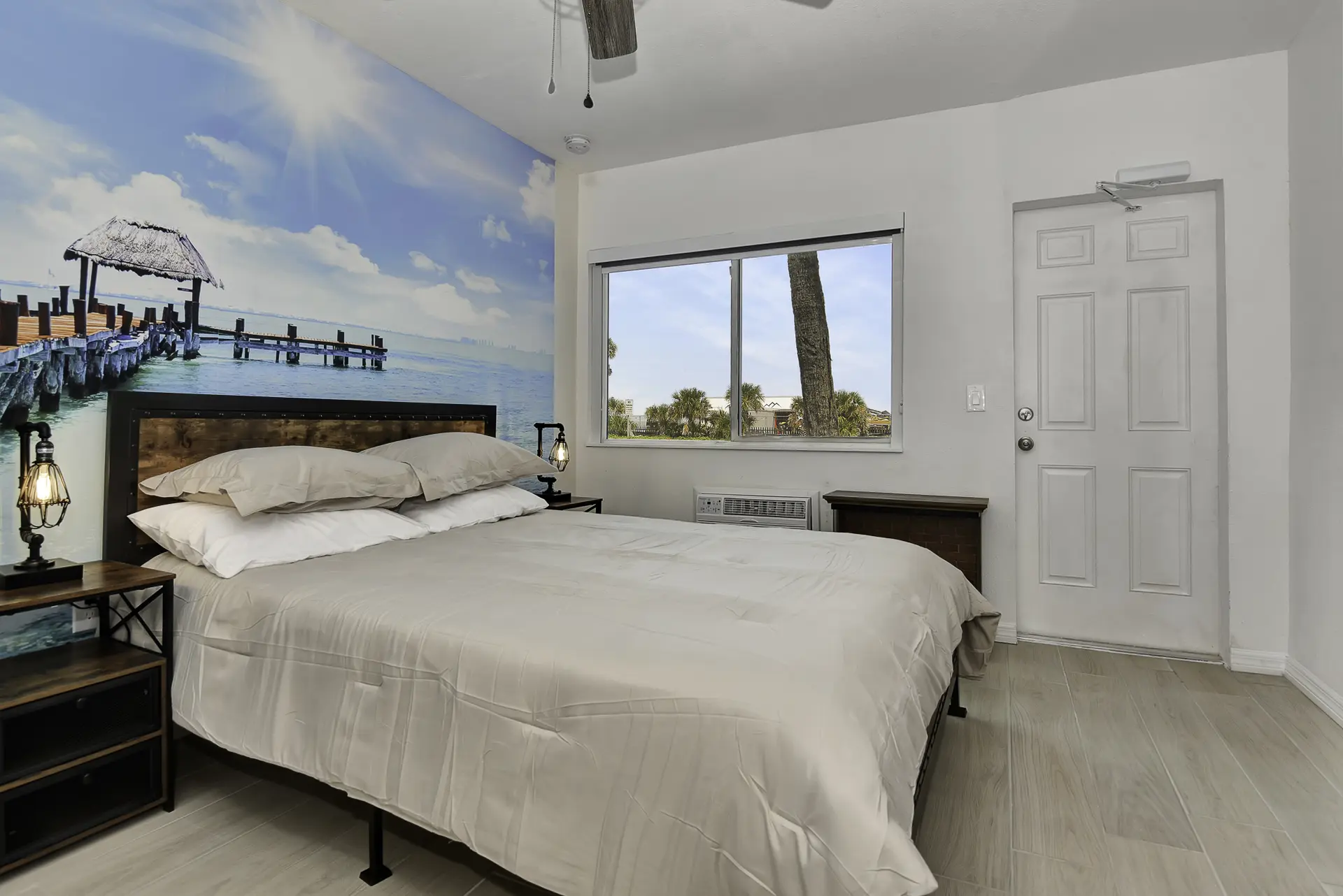 A bedroom with a bed, a window with a view of palm trees, and a door.