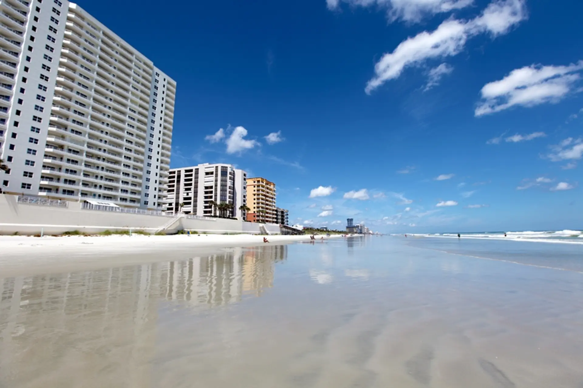 A bright, wide beach with a clear blue sky and white clouds, reflecting buildings.