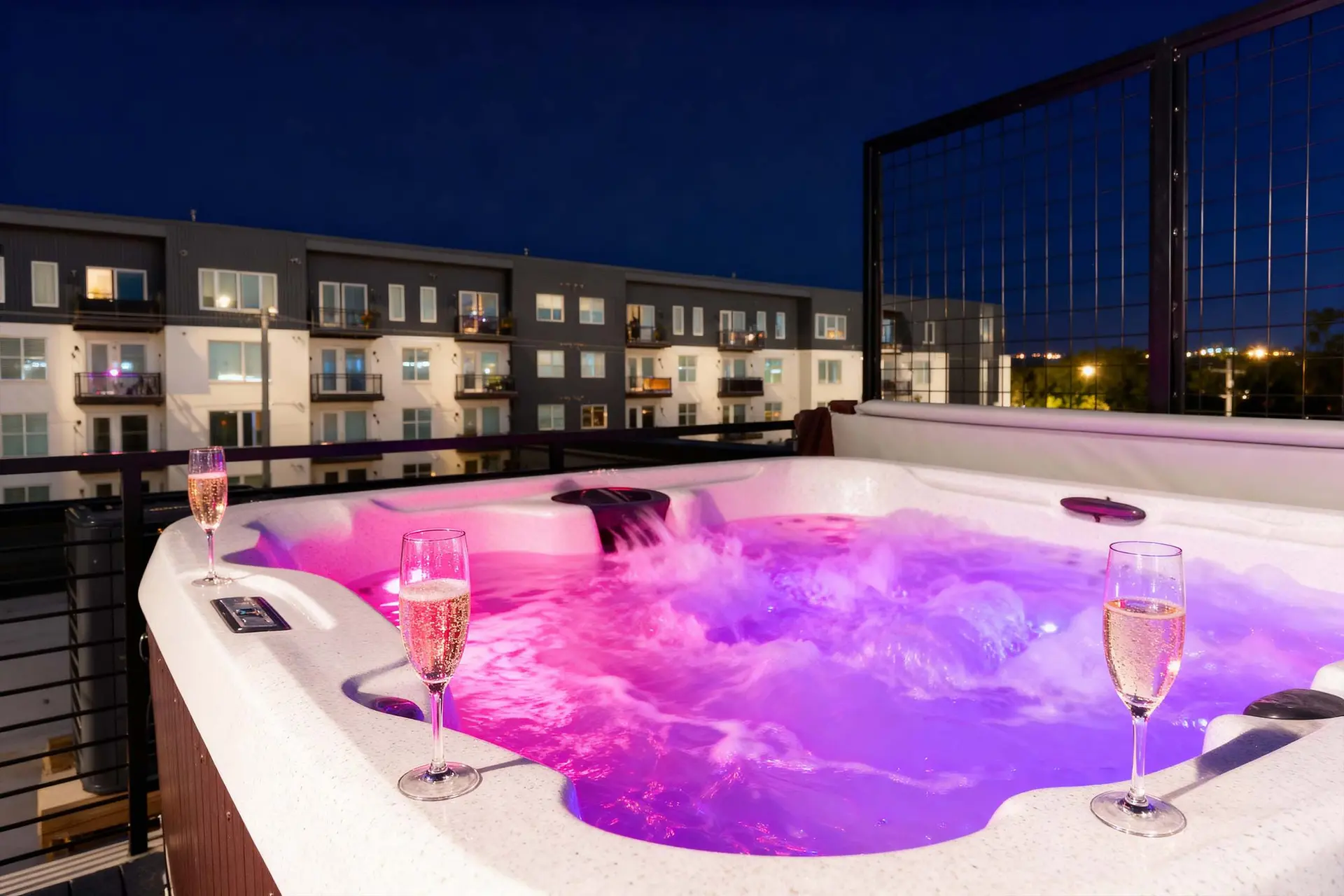 A hot tub with champagne glasses sits on a balcony at night, with a building in the background.