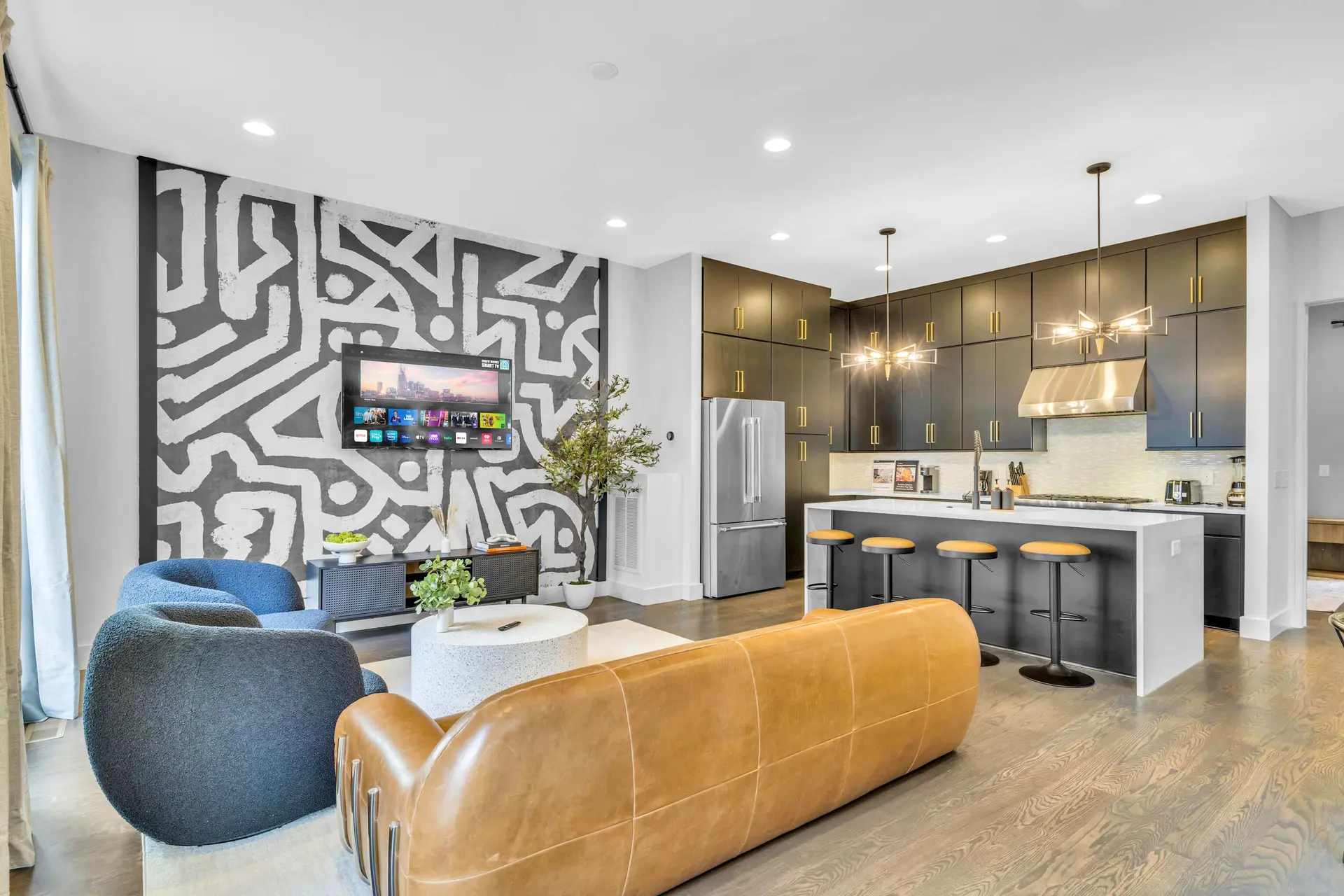 A modern living room and kitchen with a gray abstract mural, tan sofa, and blue chair.