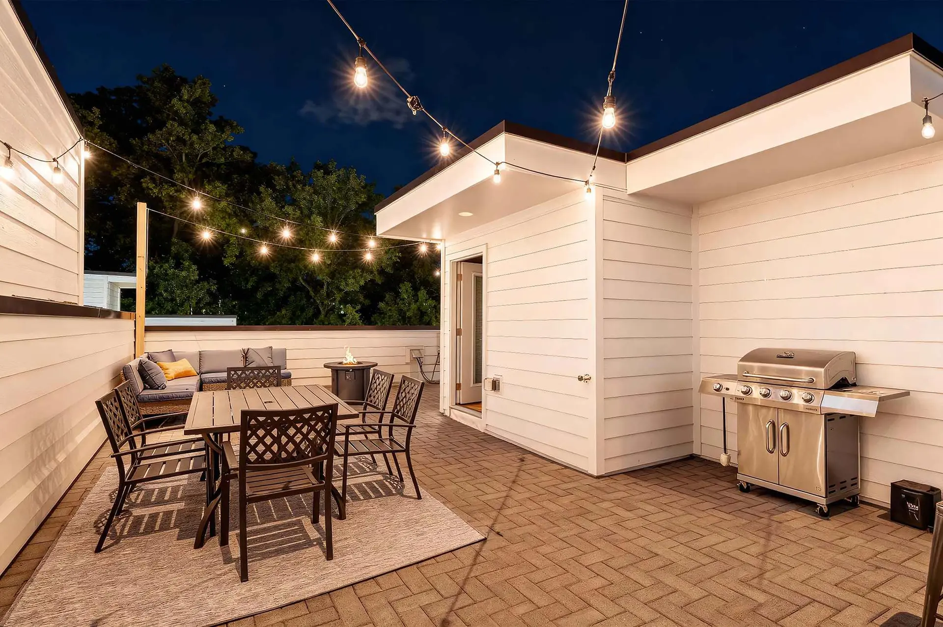 Outdoor patio at night with dining set, grill, and string lights.