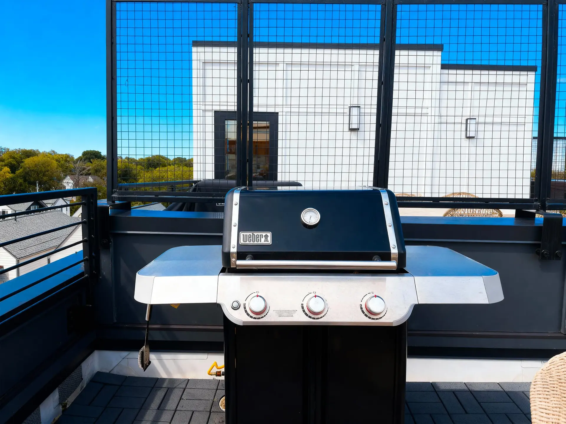 A Weber gas grill sits on a rooftop patio with a modern building and trees in the background.