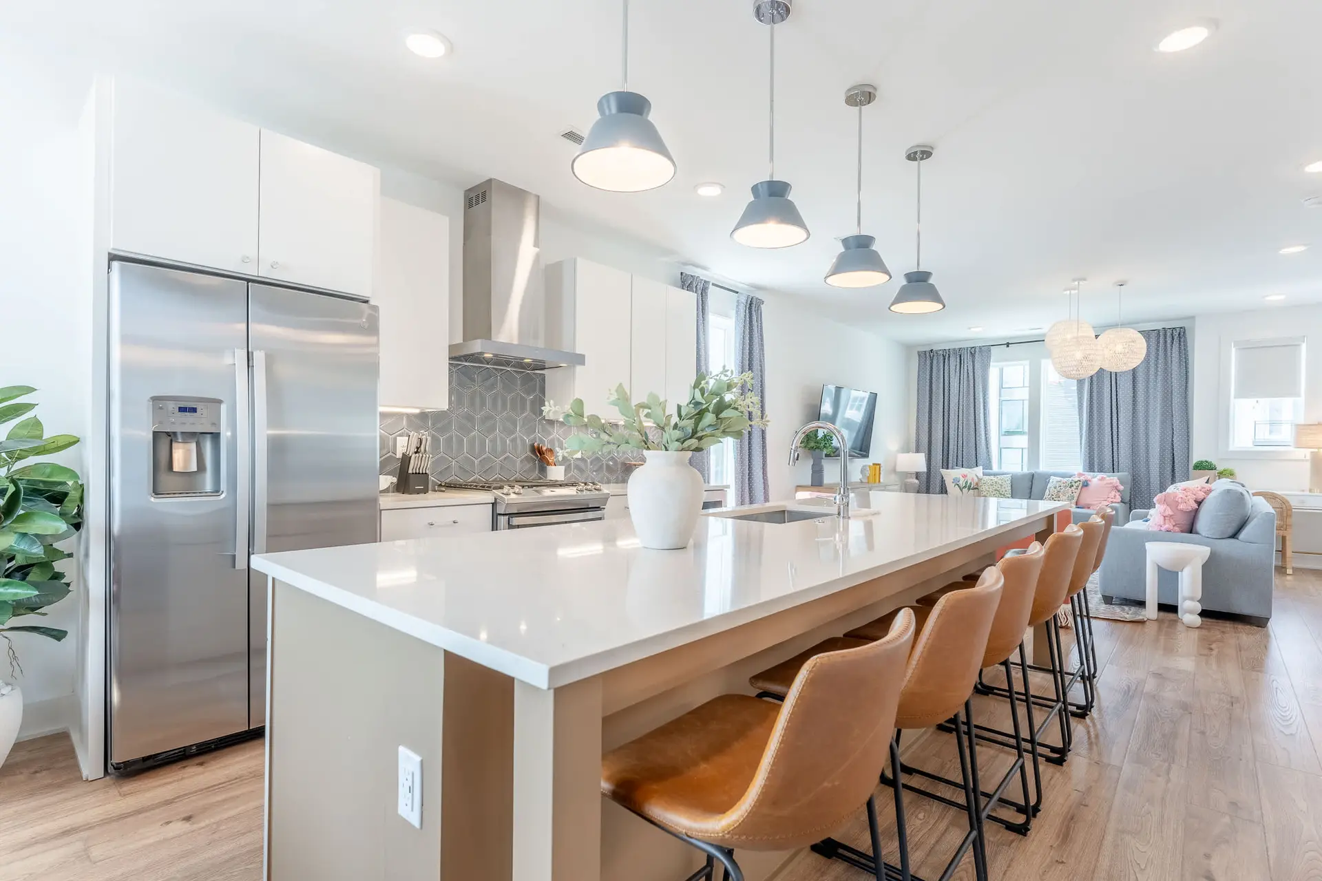 A modern kitchen island with bar stools faces a living room with blue sofas and decorative pillows.