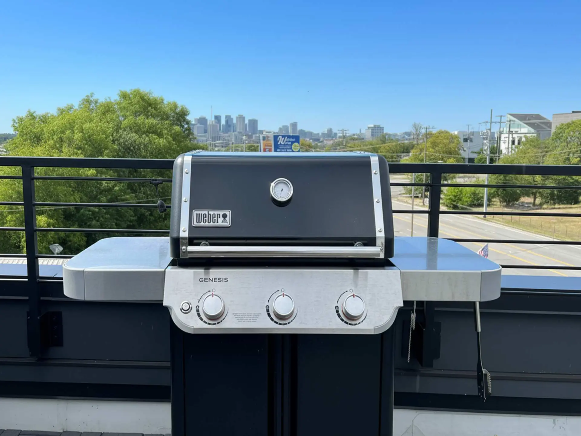A Weber Genesis grill sits on a balcony with a city skyline in the distance.