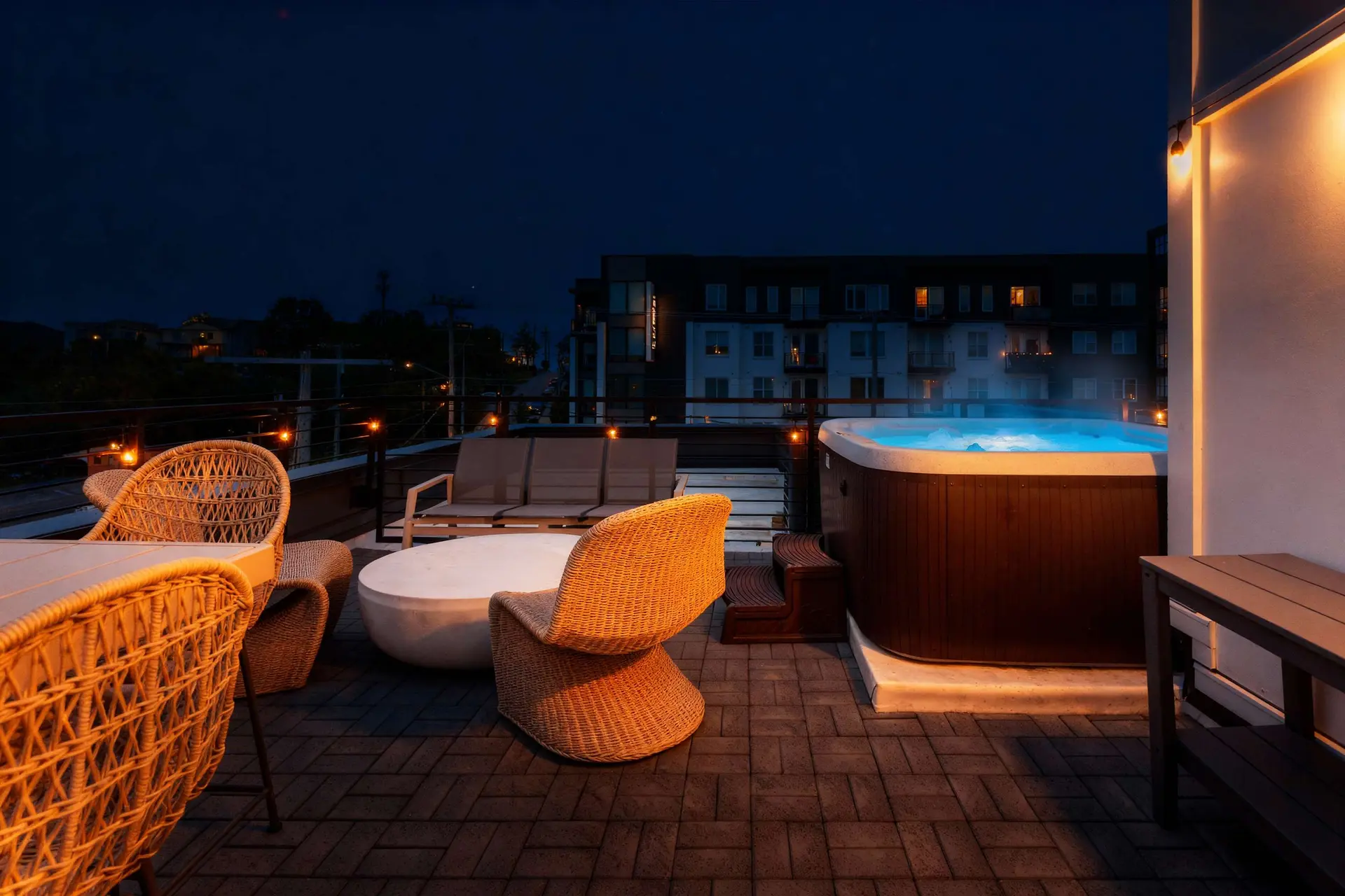 A rooftop patio at night with a hot tub, wicker chairs, a table, and a modern apartment building in 