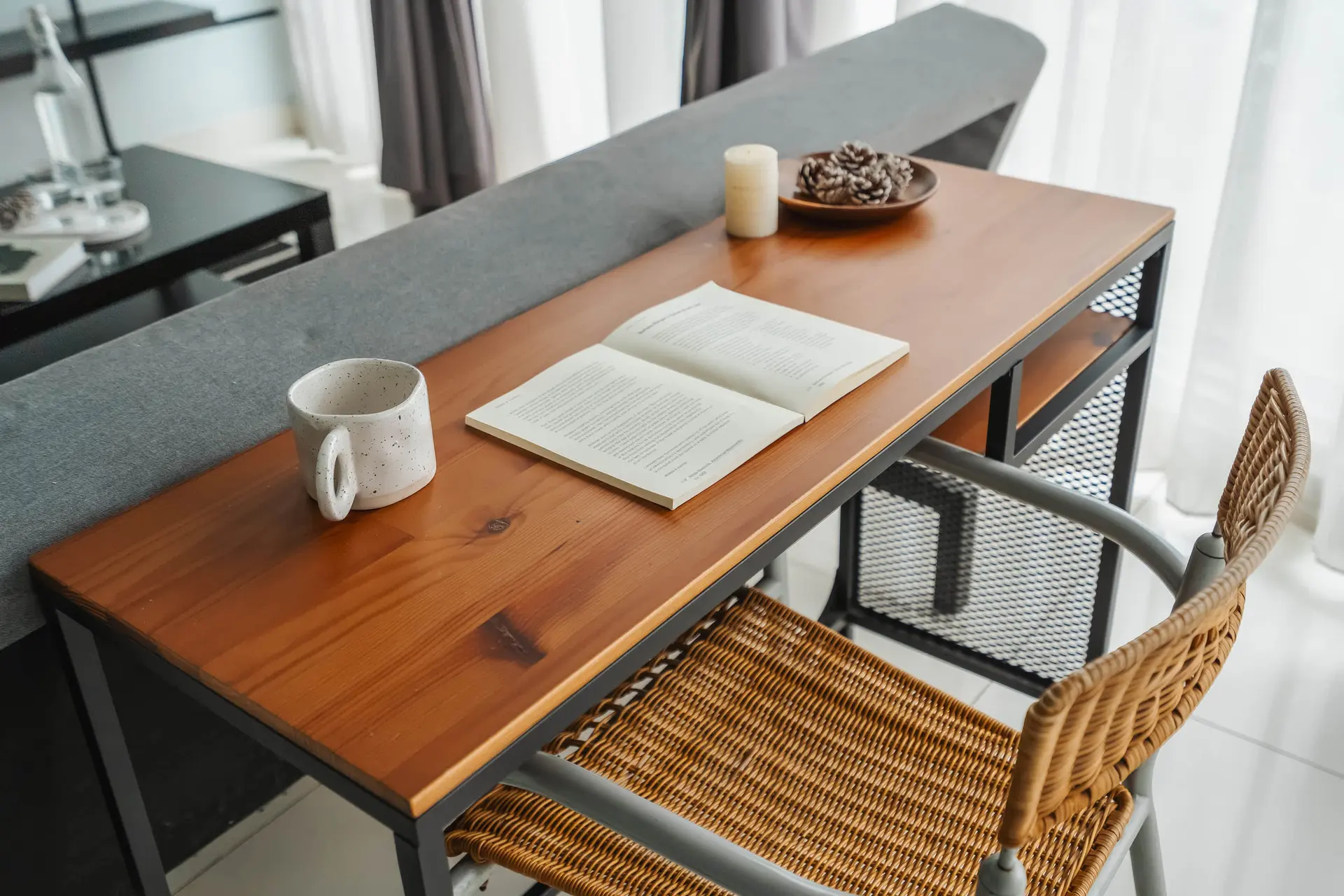 A mug, an open book, a candle, and pinecones sit on a wooden desk.