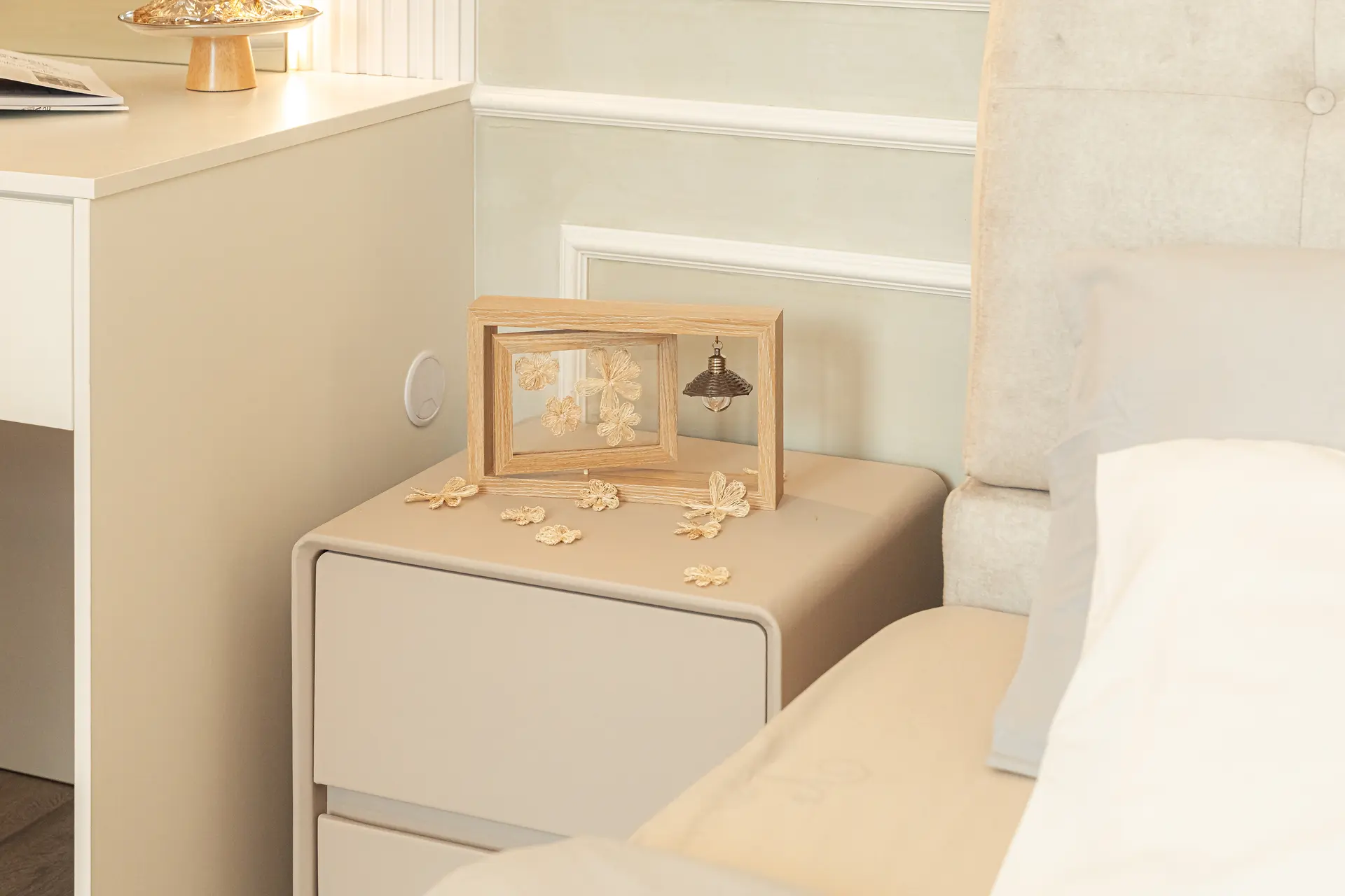A light wood picture frame with woven straw flowers and a tiny lamp is placed on a beige nightstand.