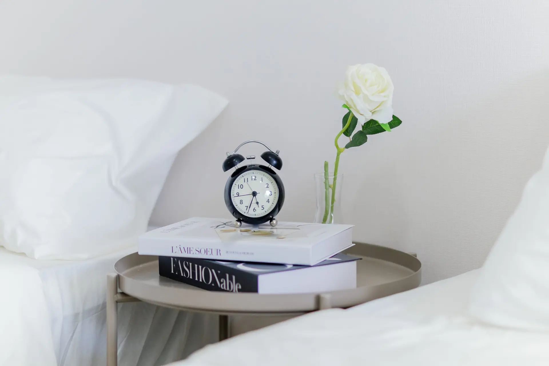 A black alarm clock rests on a stack of books next to a white rose in a vase on a bedside table.