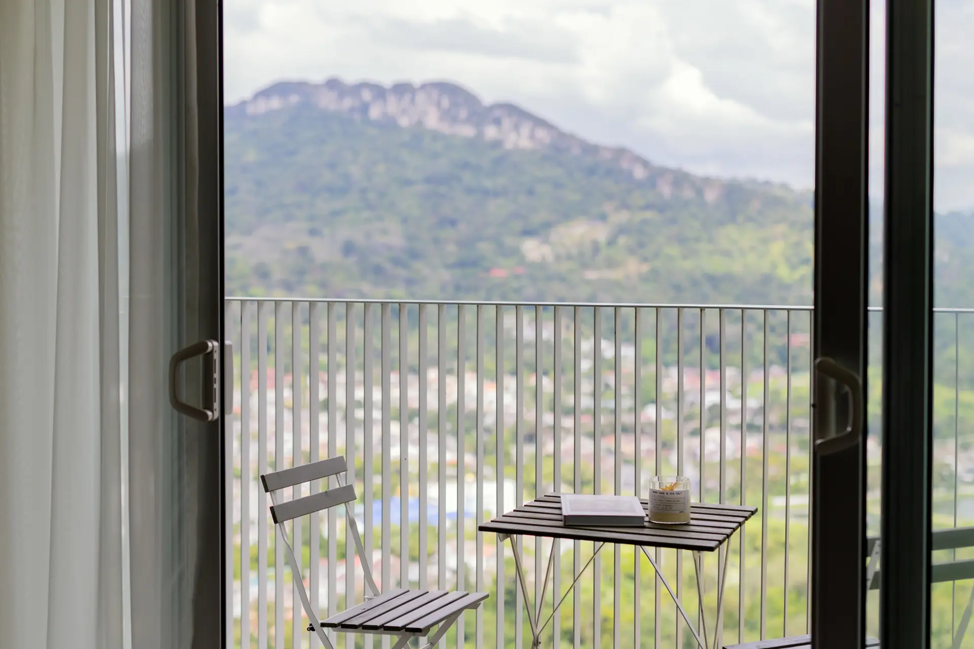 A balcony with a chair and table overlooks a city and mountain view.