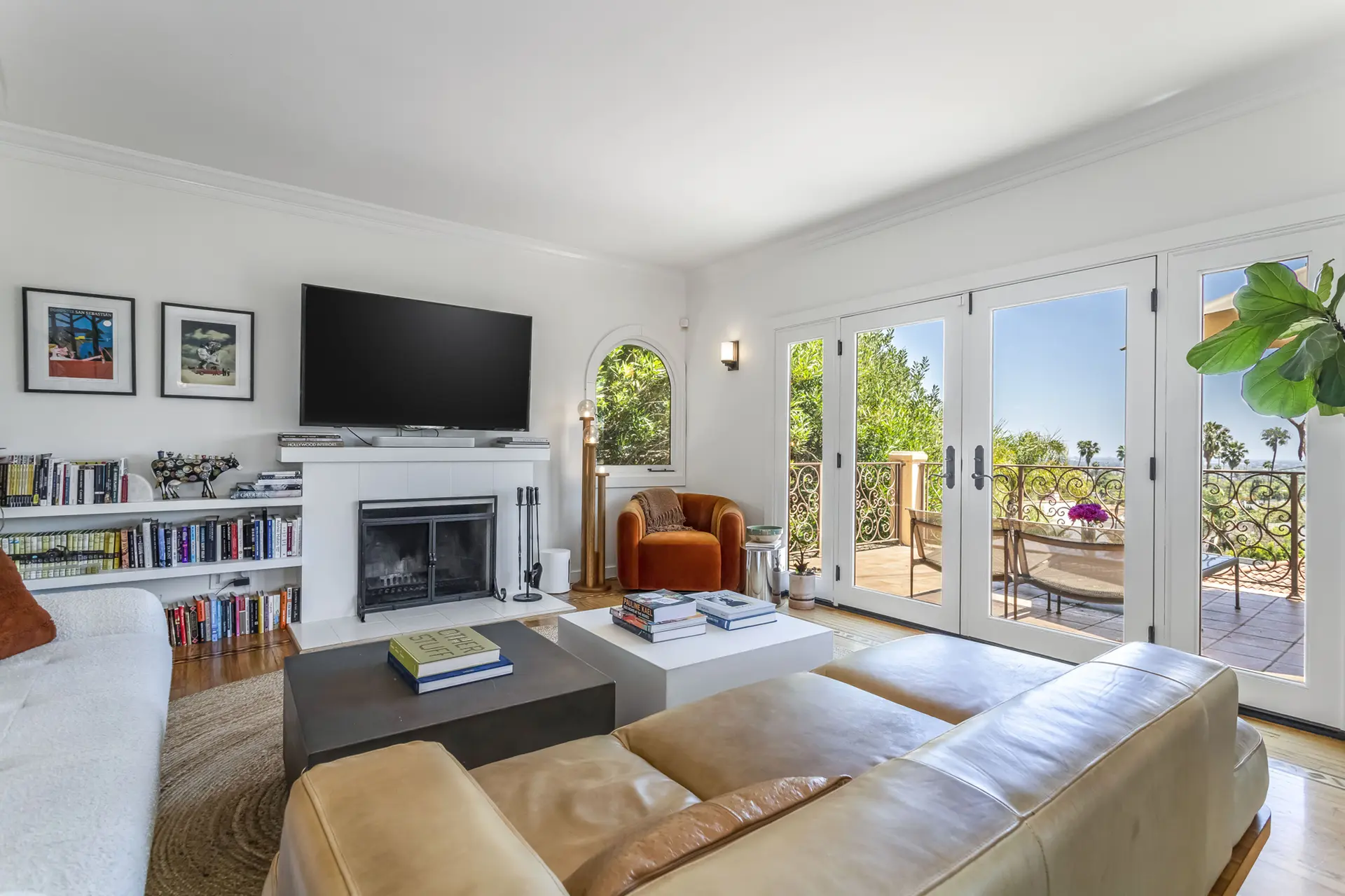 A living room with a fireplace, TV, and large windows overlooking a balcony with palm trees.