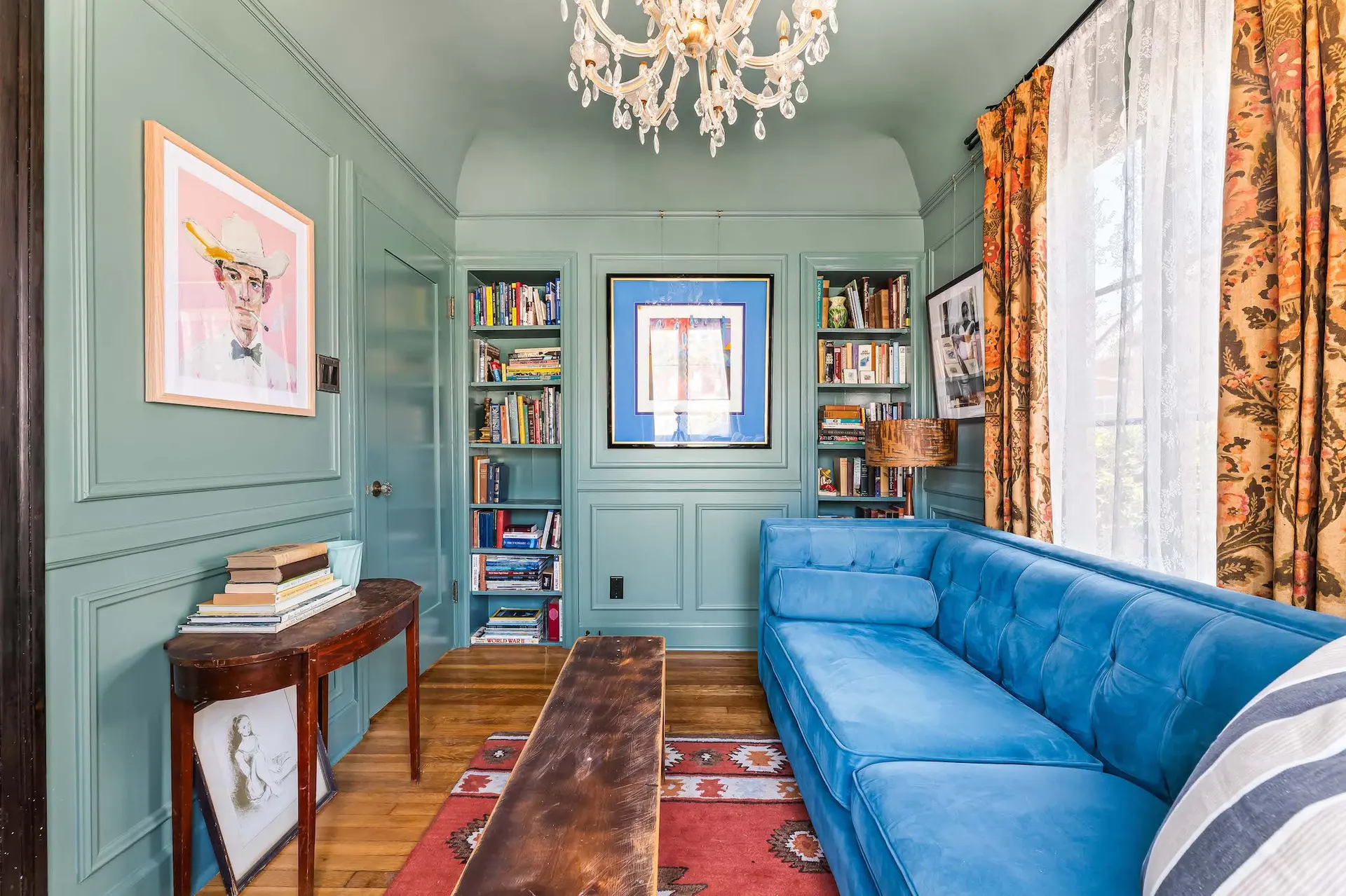 A blue velvet sofa sits in front of a bookshelf, a chandelier hangs above.