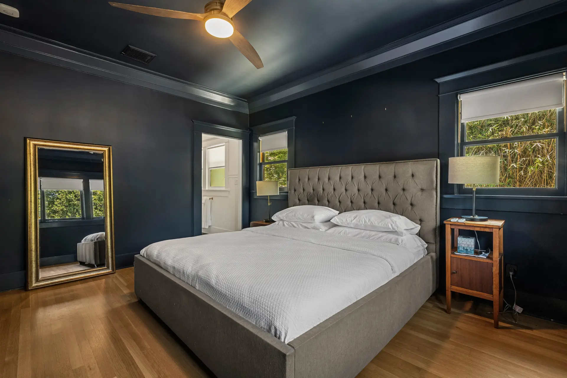 Bedroom with dark blue walls, a large tufted bed, a gold mirror, and a wooden nightstand.