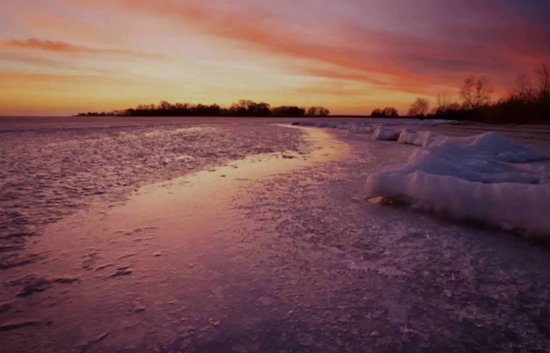 A frozen lake reflects a dramatic sunset with orange and pink hues.