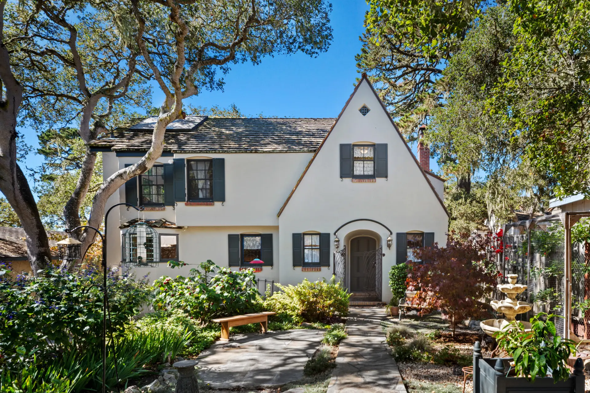 Charming stucco house with dark shutters, surrounded by lush greenery and mature trees.
