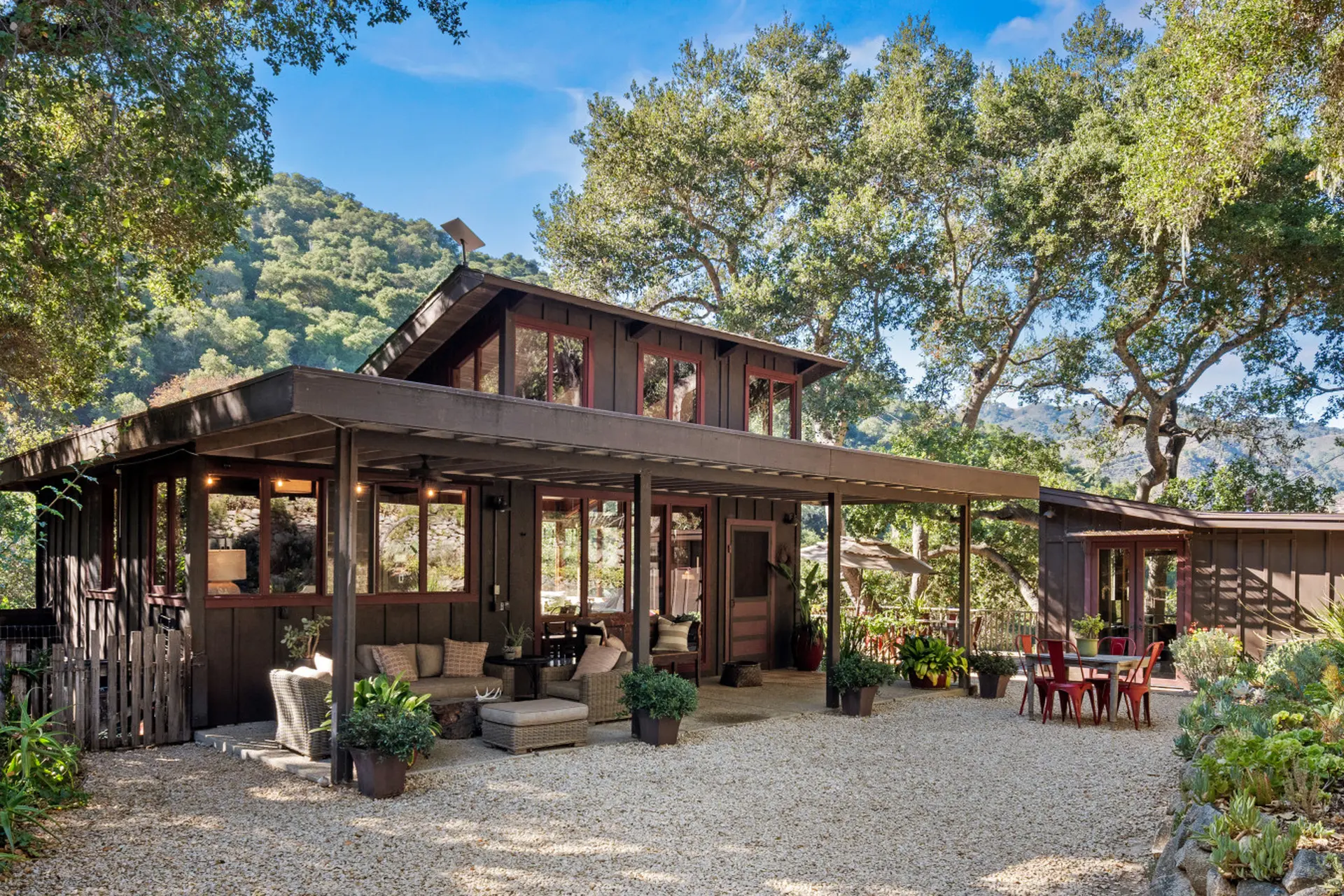 Brown wooden house with a covered porch, outdoor furniture, and trees in the background.