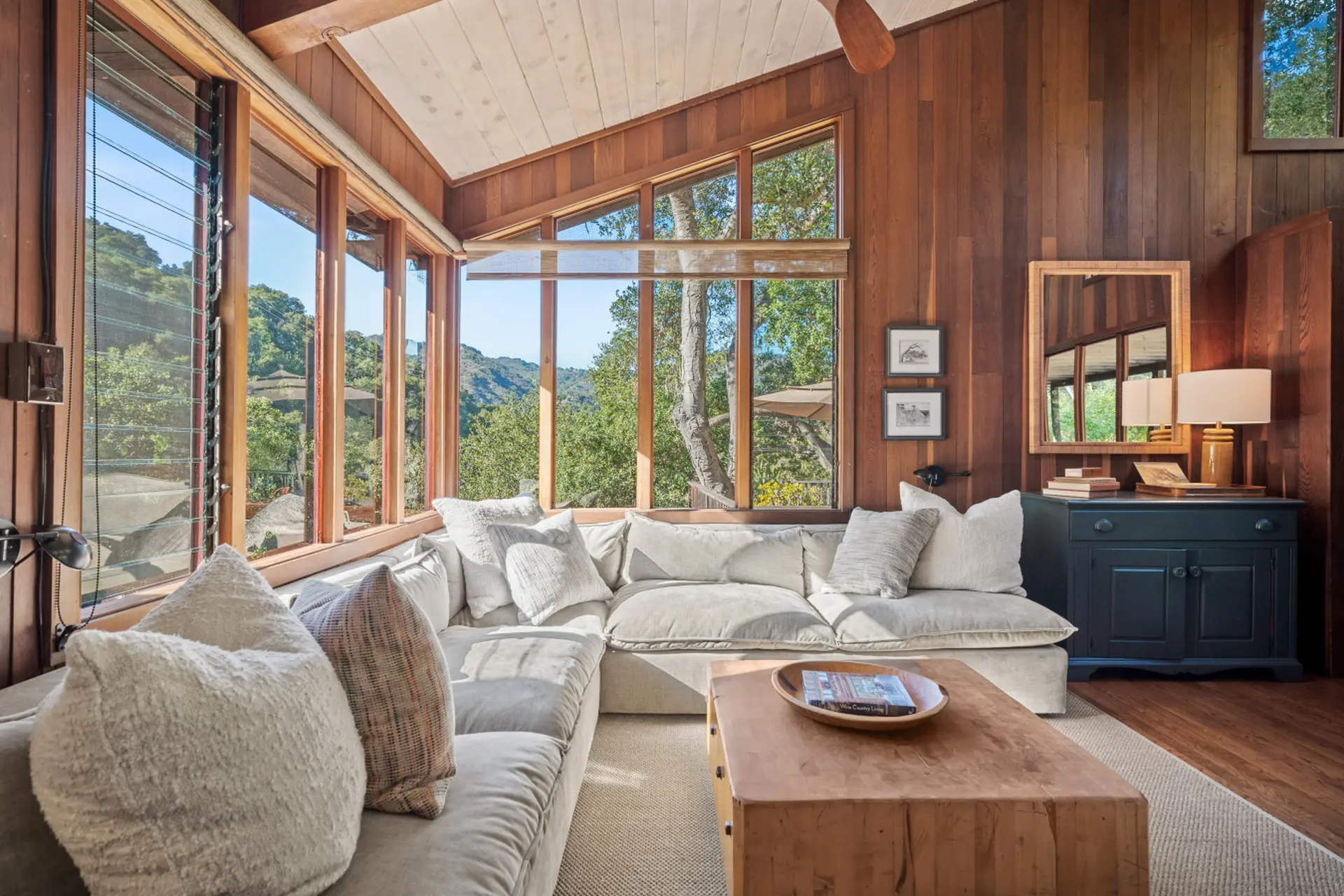 Cozy living room with wood paneling, large sectional sofa, and windows with views of trees.