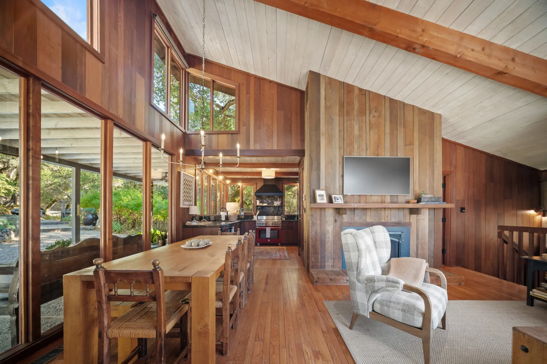 A dining area and kitchen with wood paneling and large windows overlook a lush green landscape.