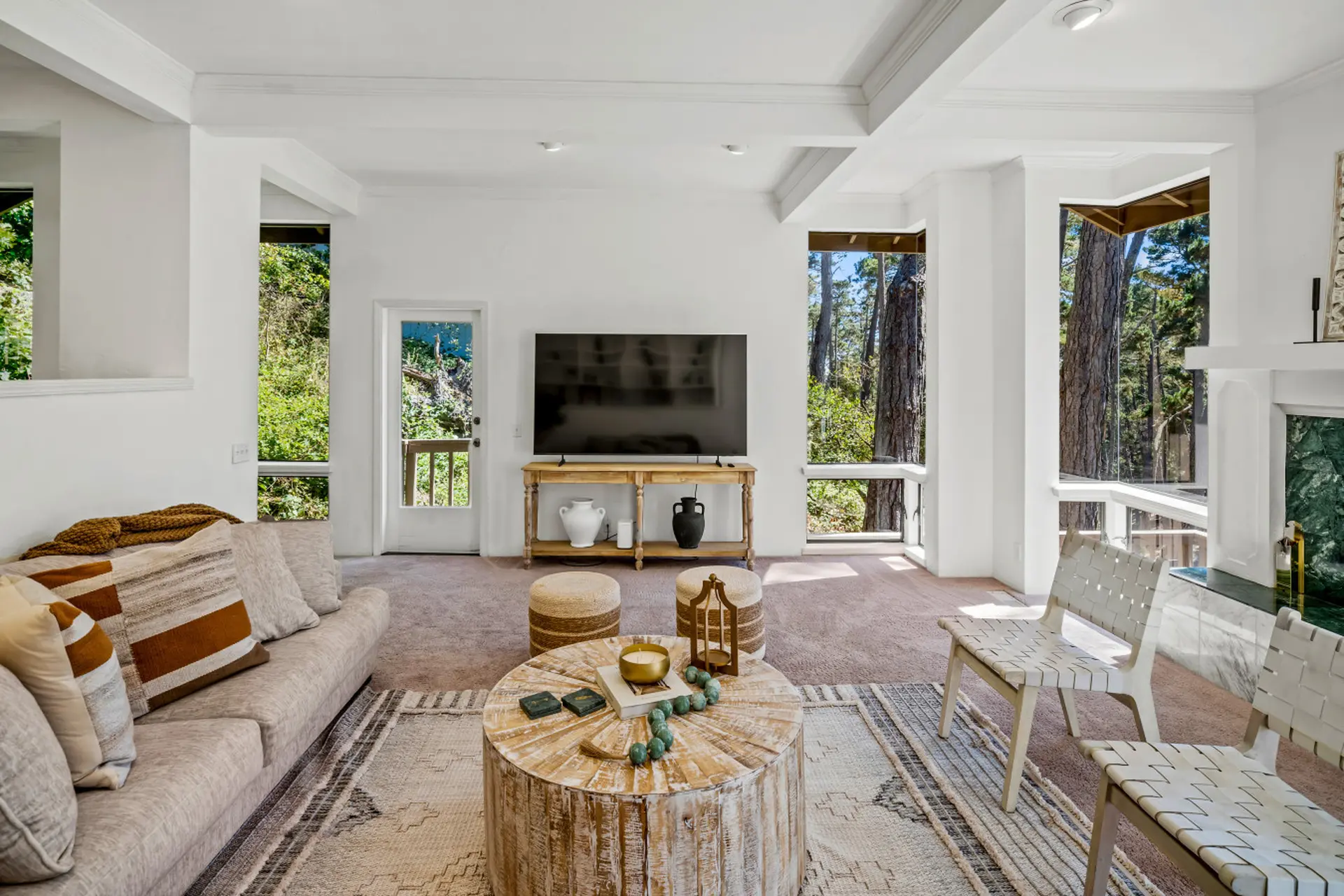 A light-filled living room with a sofa, coffee table, and large windows revealing a forest view.