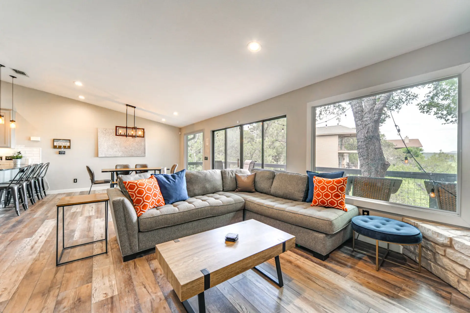 A modern living room with a gray sectional couch, wooden floors, and large windows overlooking a woo
