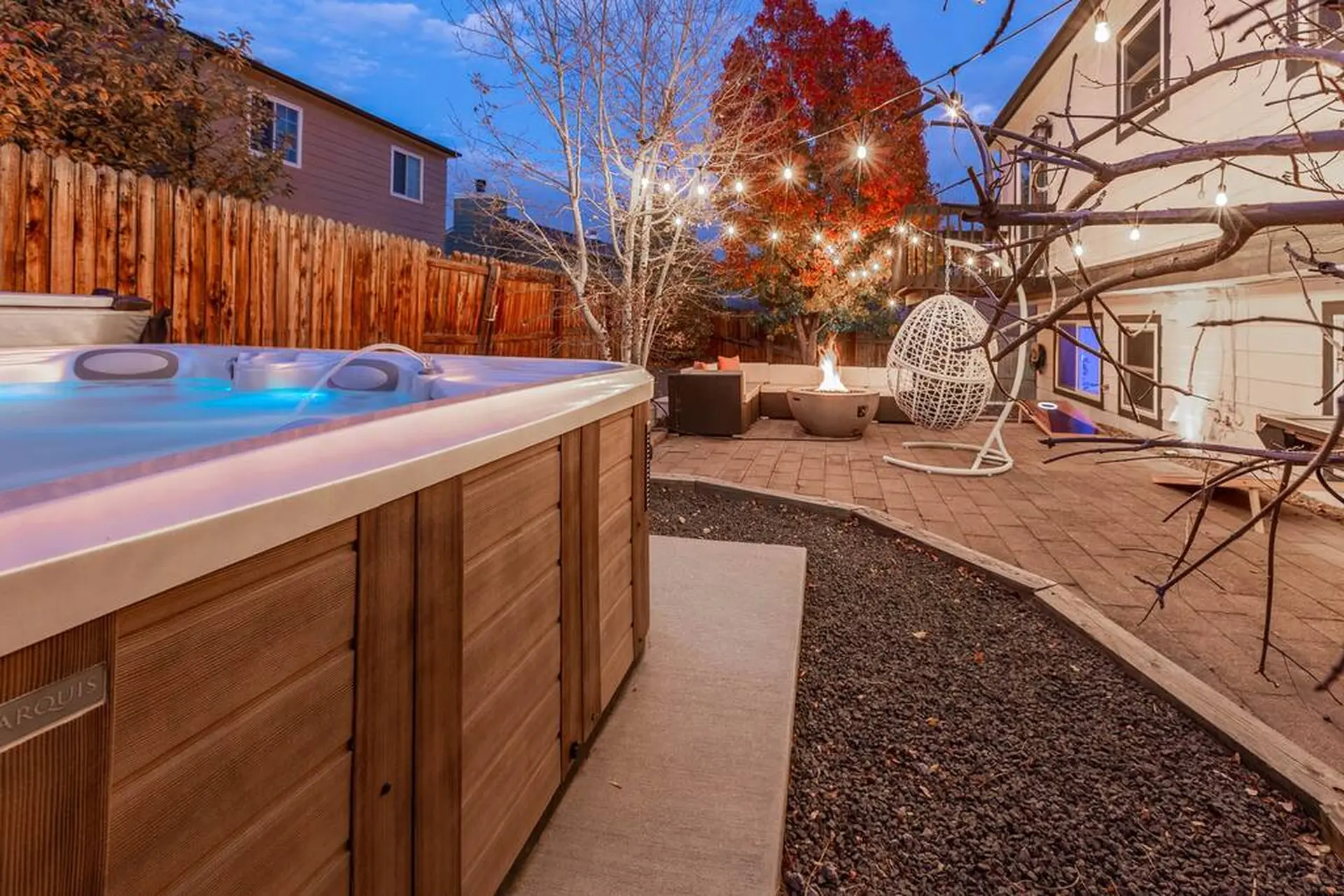 A hot tub sits in a backyard patio with a fire pit and string lights.