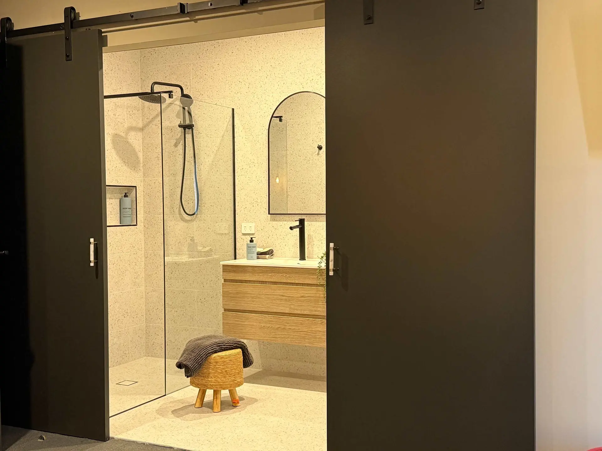 A bathroom with a shower, sink, and wooden vanity, accessed through a sliding door.