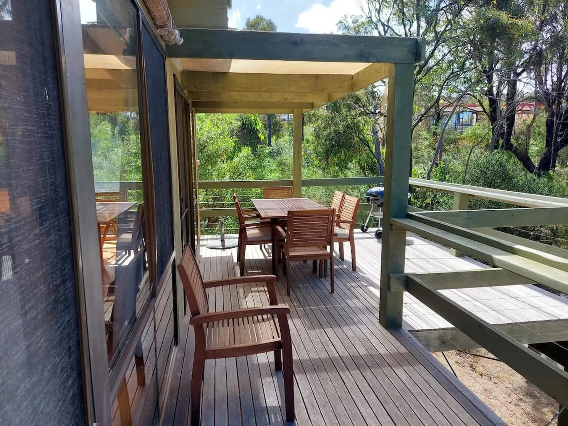 Wooden deck with chairs and tables, a BBQ, and a view of trees and houses.