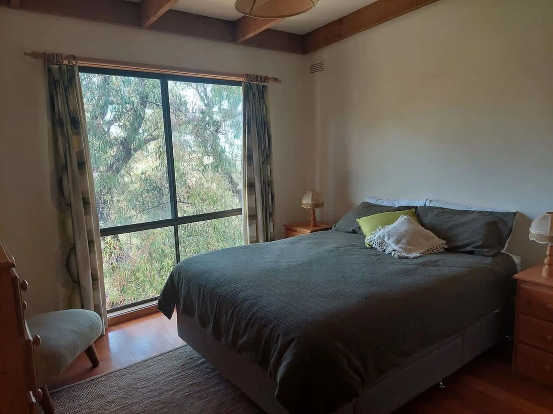 Bedroom with large window and trees, bed with green and white pillows, bedside tables and lamps.