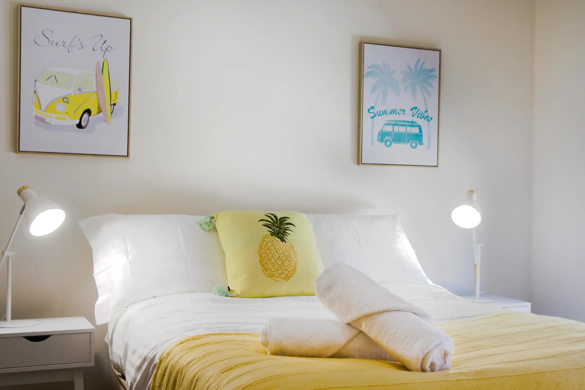 A brightly lit bedroom features a bed with white linens, a yellow pineapple pillow, and two rolled w