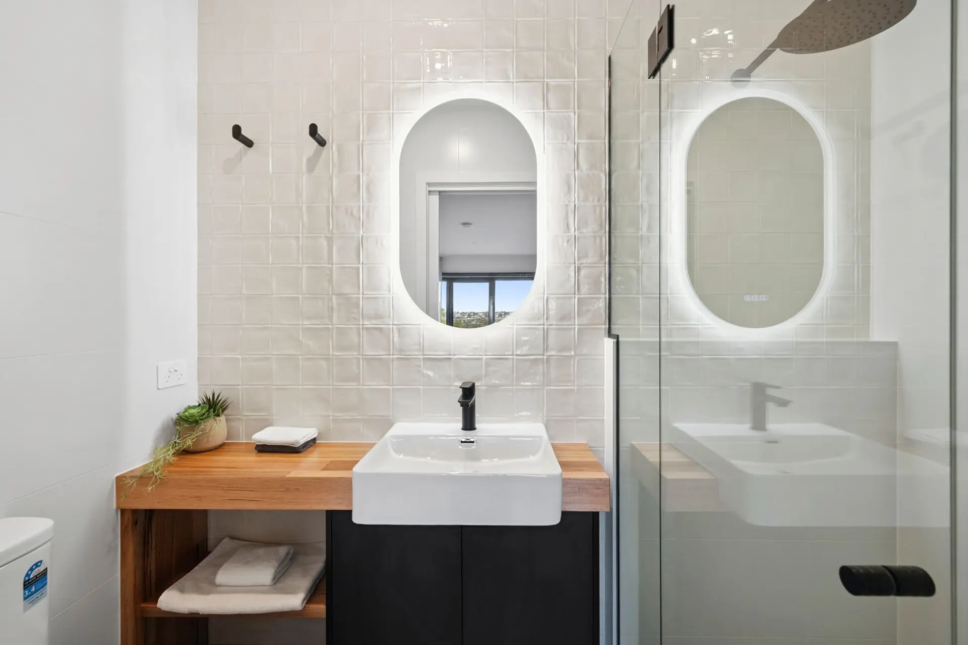 Modern bathroom with a wooden vanity, black cabinets, white square tile, and an oval illuminated mir