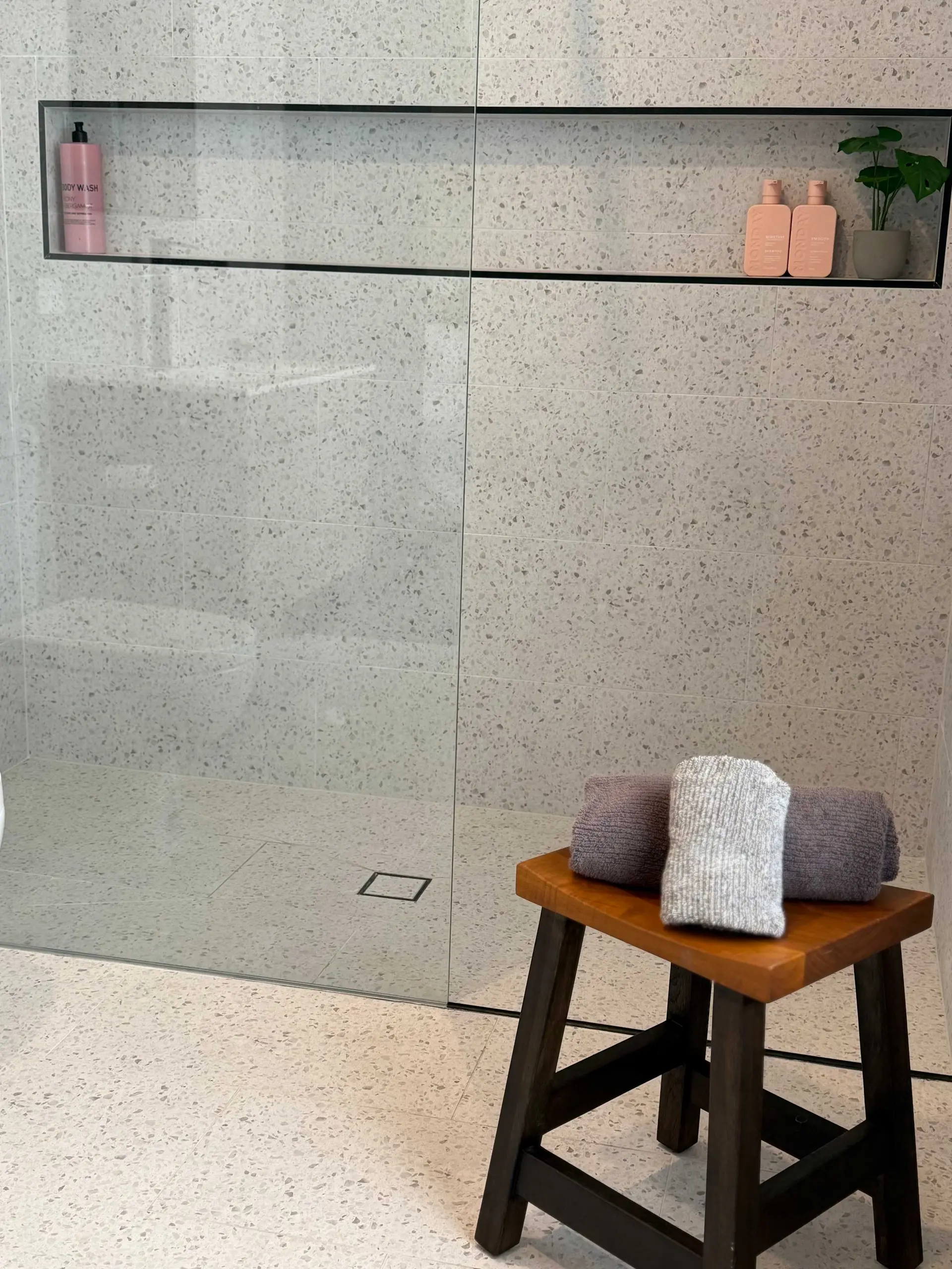 A wooden stool with towels sits in a bathroom with speckled gray tiles and a glass shower.
