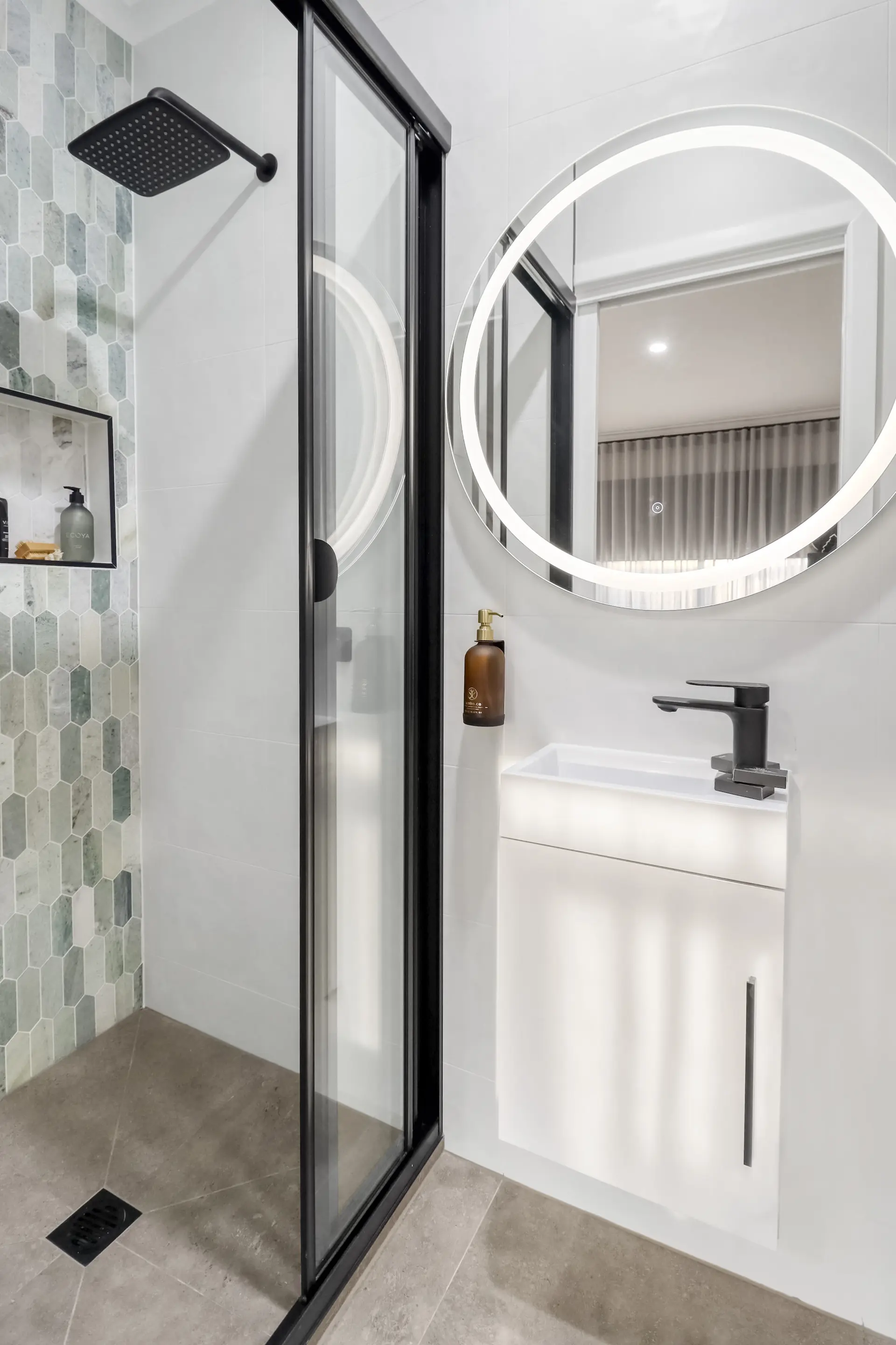 Modern bathroom with a square black showerhead, glass shower door, and a lit round mirror above a wh