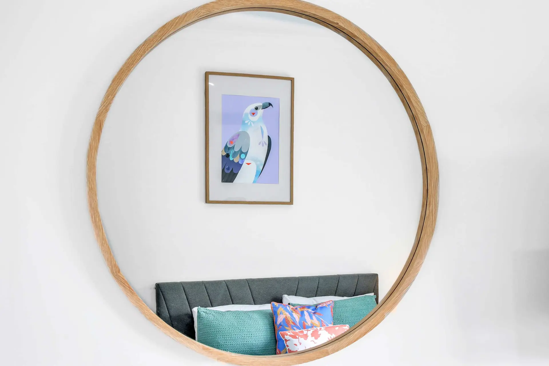 A large round mirror reflects a painting of a bird and a bed with pillows.