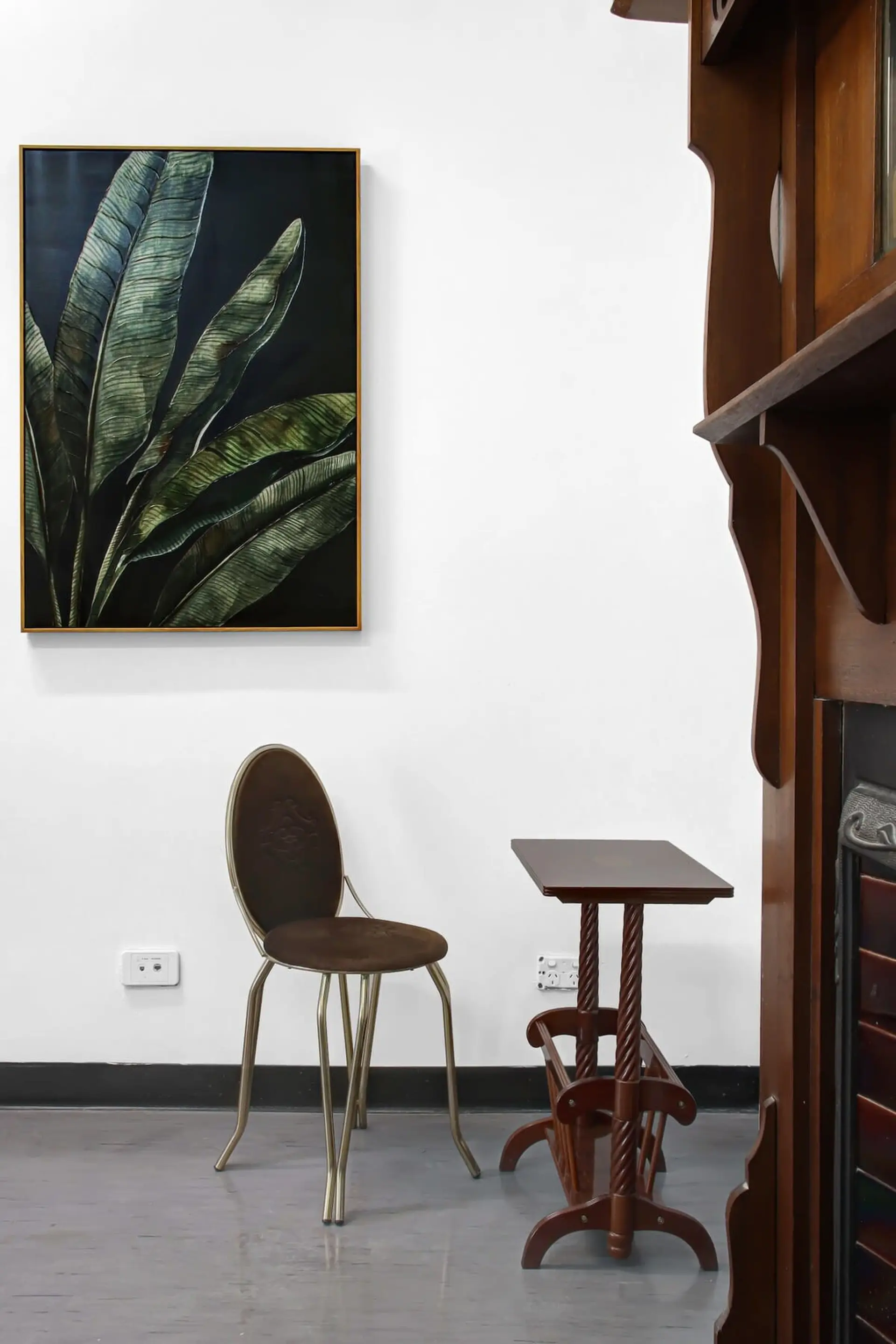 A painting of green leaves hangs above a gold-legged chair and a wooden side table.