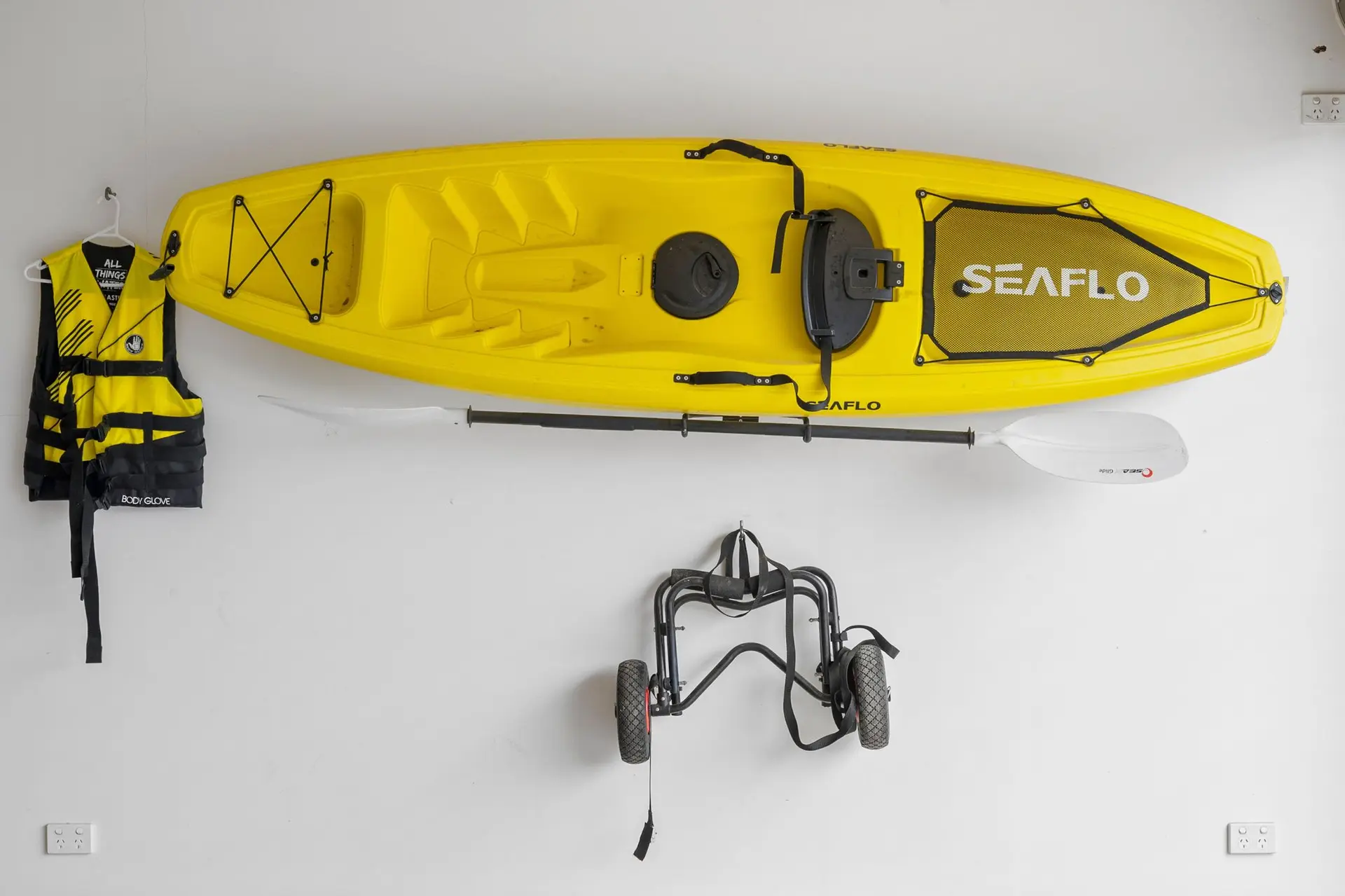 A yellow kayak and paddle are mounted on a wall, with a life vest and kayak cart below.