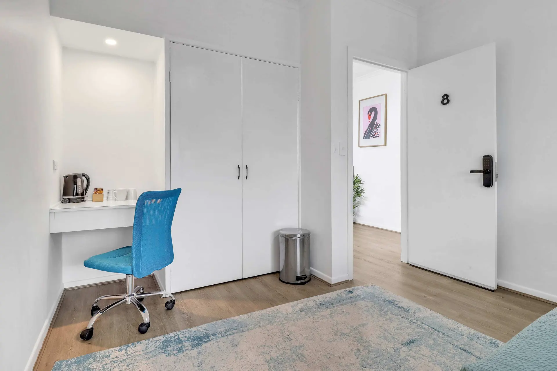 A blue desk chair sits by a white desk with a kettle and cups. A door with the number 8 hangs open, 