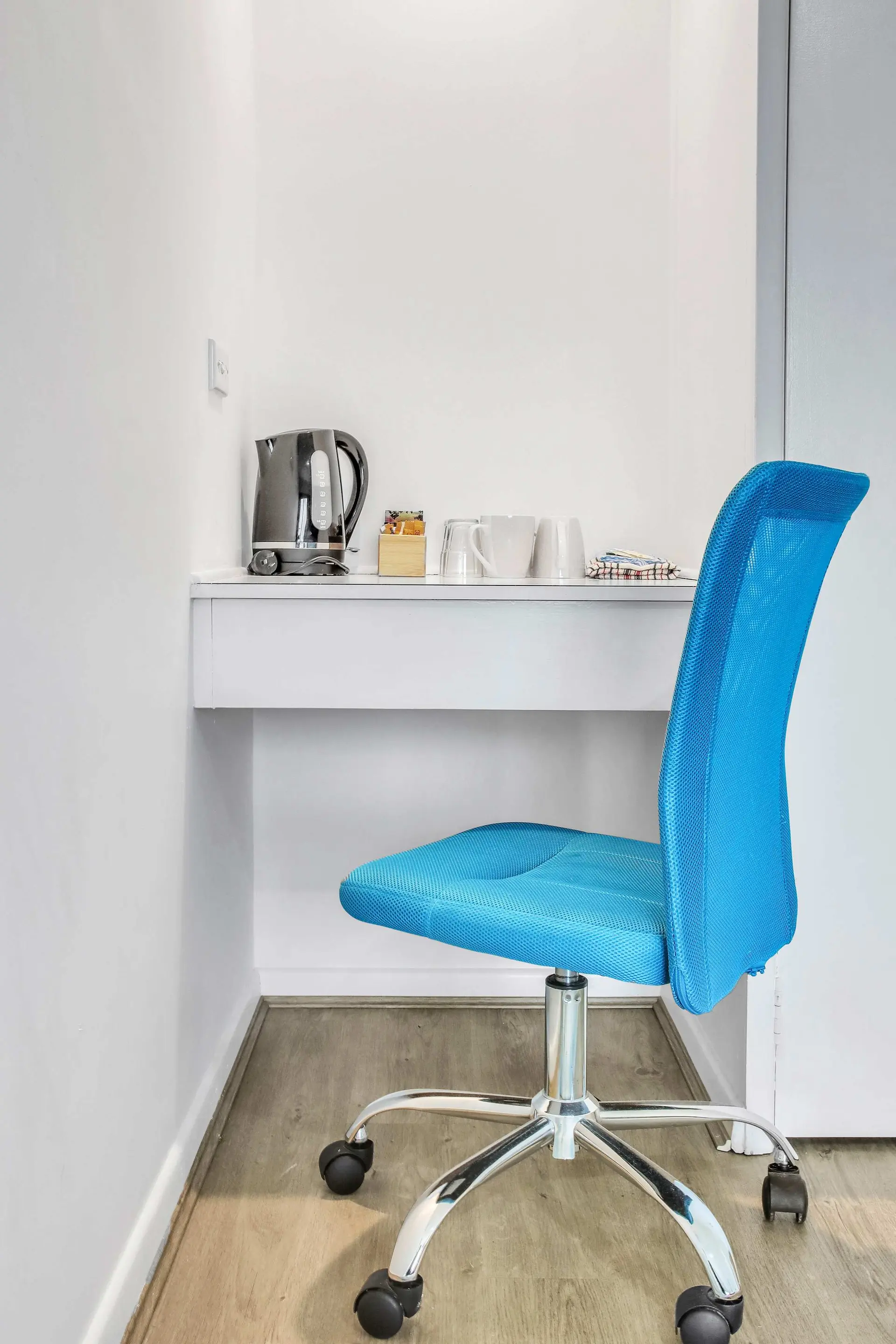 A blue mesh office chair sits in front of a white desk with a kettle and mugs.