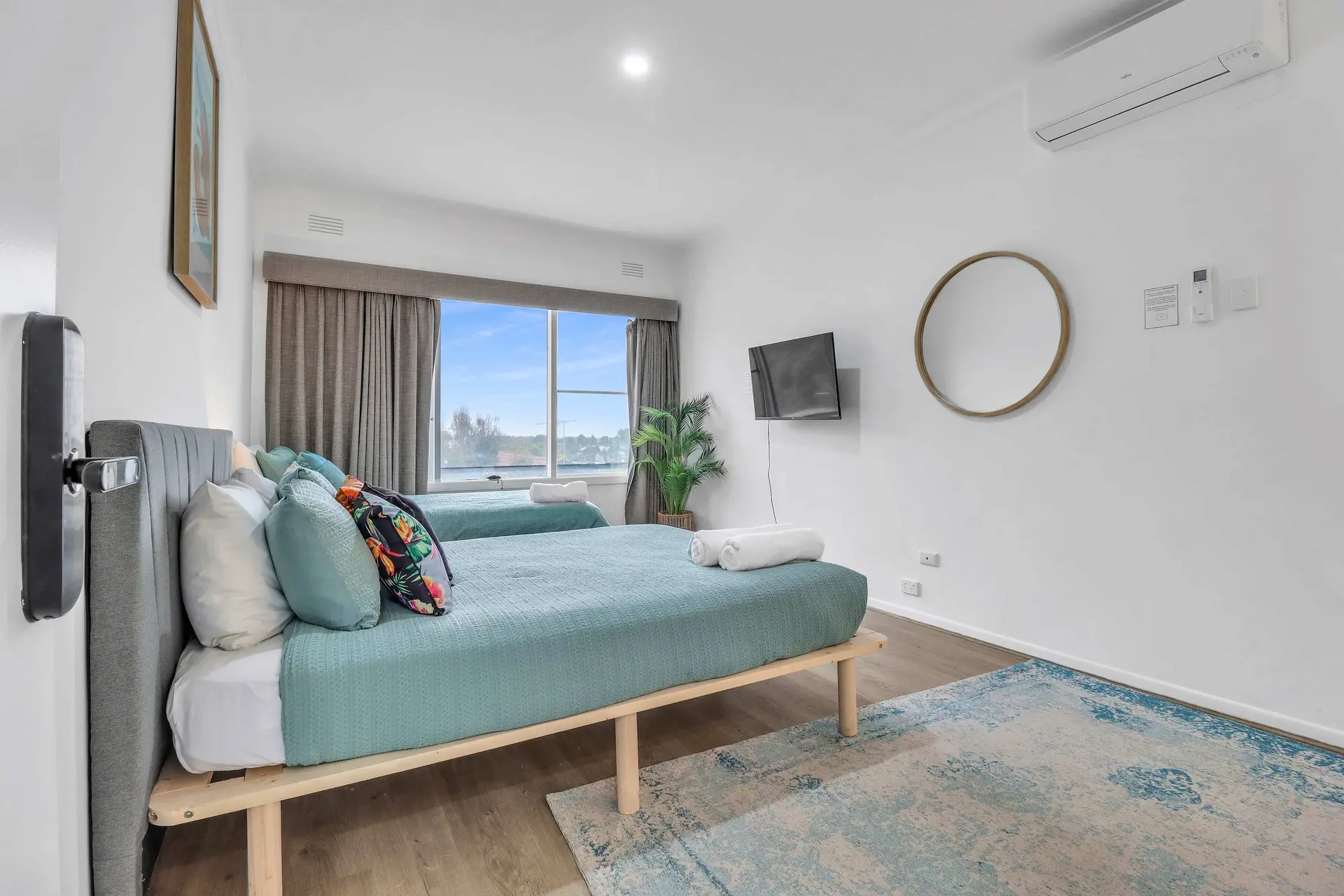 Two teal beds with plush pillows are in a room with a TV and a large window.