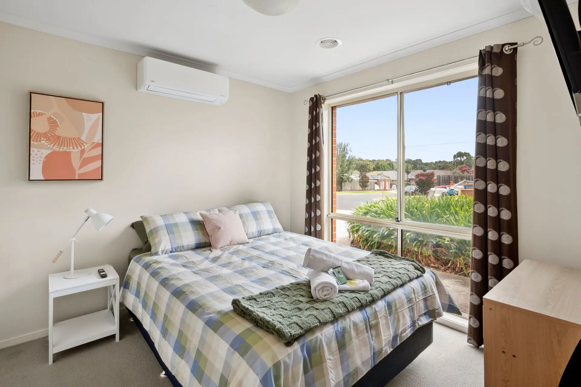A tidy bedroom features a bed with a plaid comforter, white nightstand, and a large window with brow