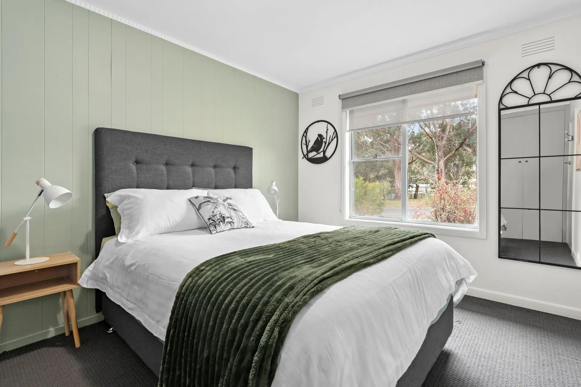 A cozy bedroom featuring a bed with white linens and a green throw, bedside table with a lamp, and a