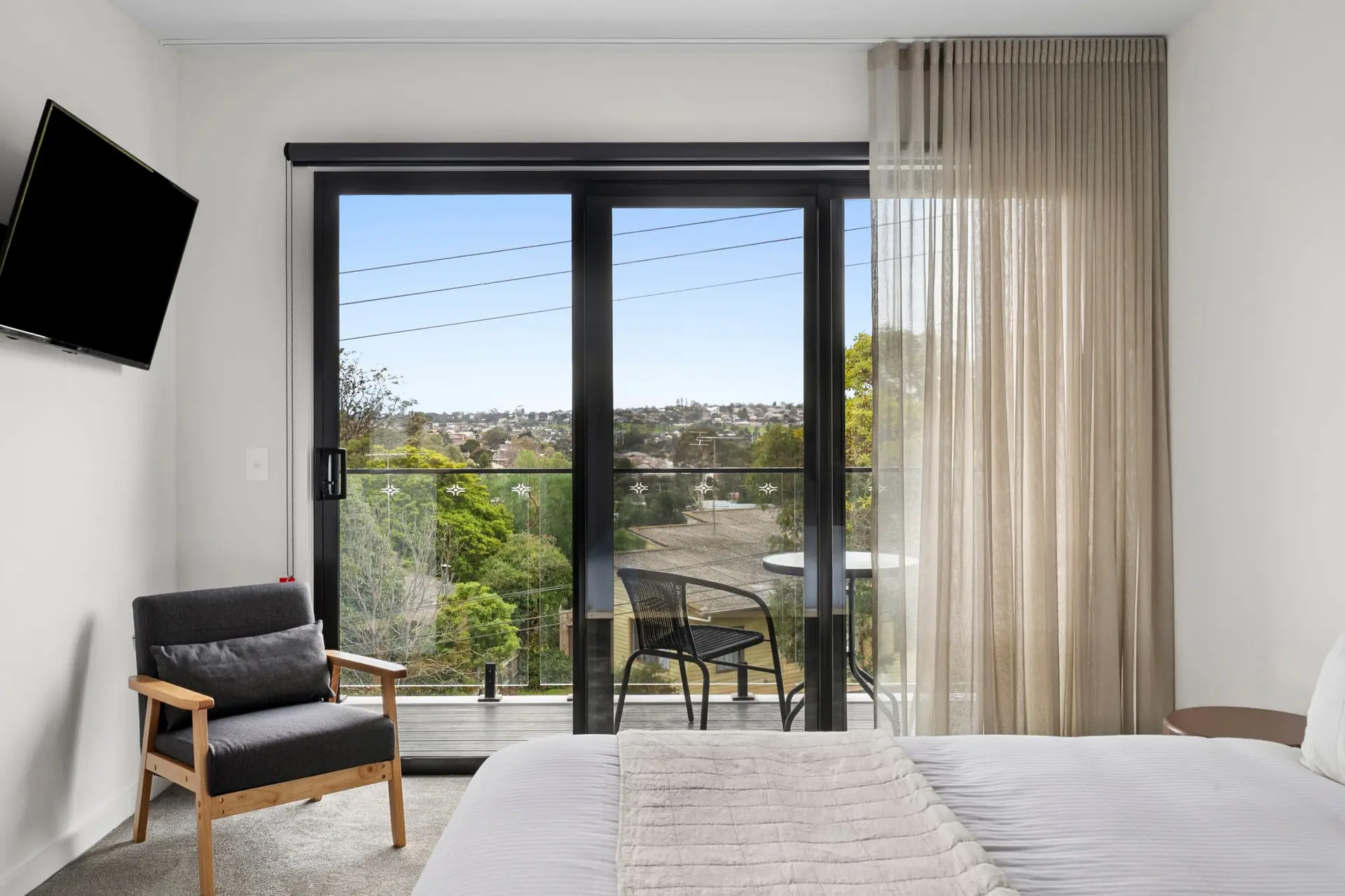 Modern bedroom with a wall-mounted TV, a plush armchair, and a large glass door opening to a balcony