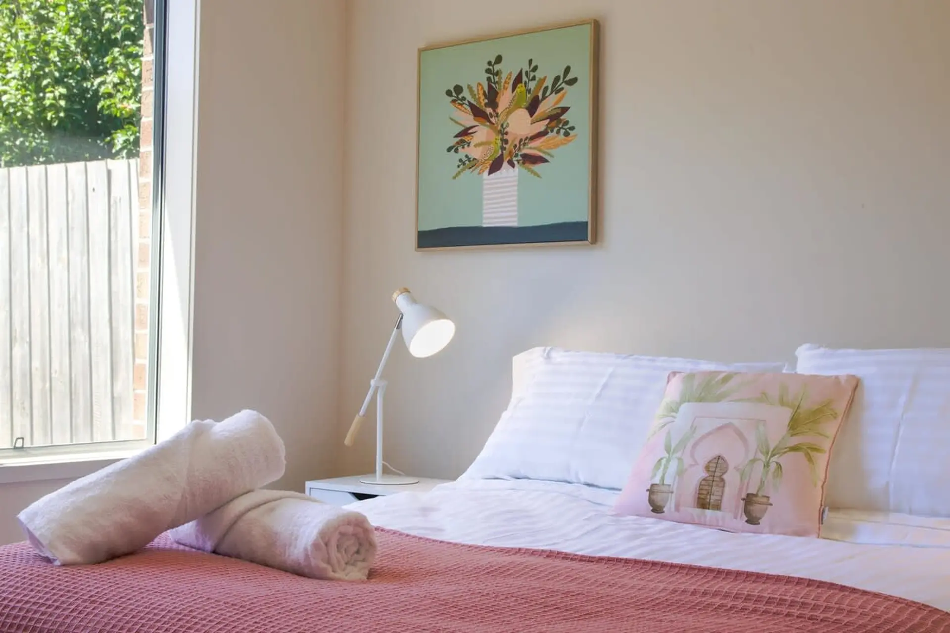 A pink bedspread with rolled towels, a white lamp, and a floral painting.