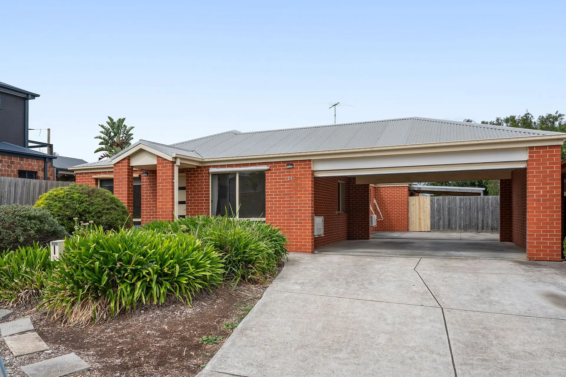 Red brick house with carport, driveway, and green bushes.
